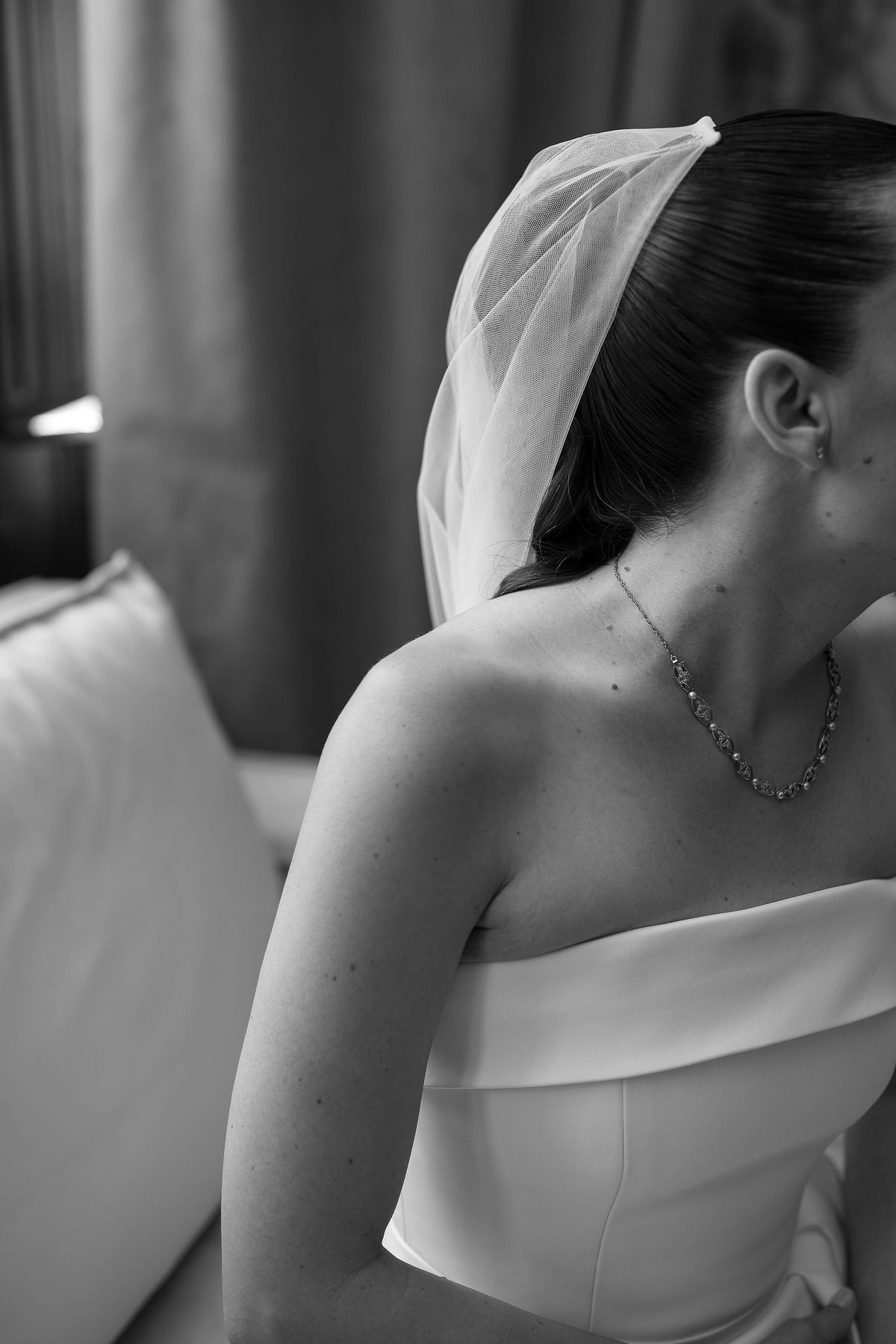 The bride, seen in profile from behind, wears a short veil, a delicate necklace and a strapless gown beside a cushioned sofa.