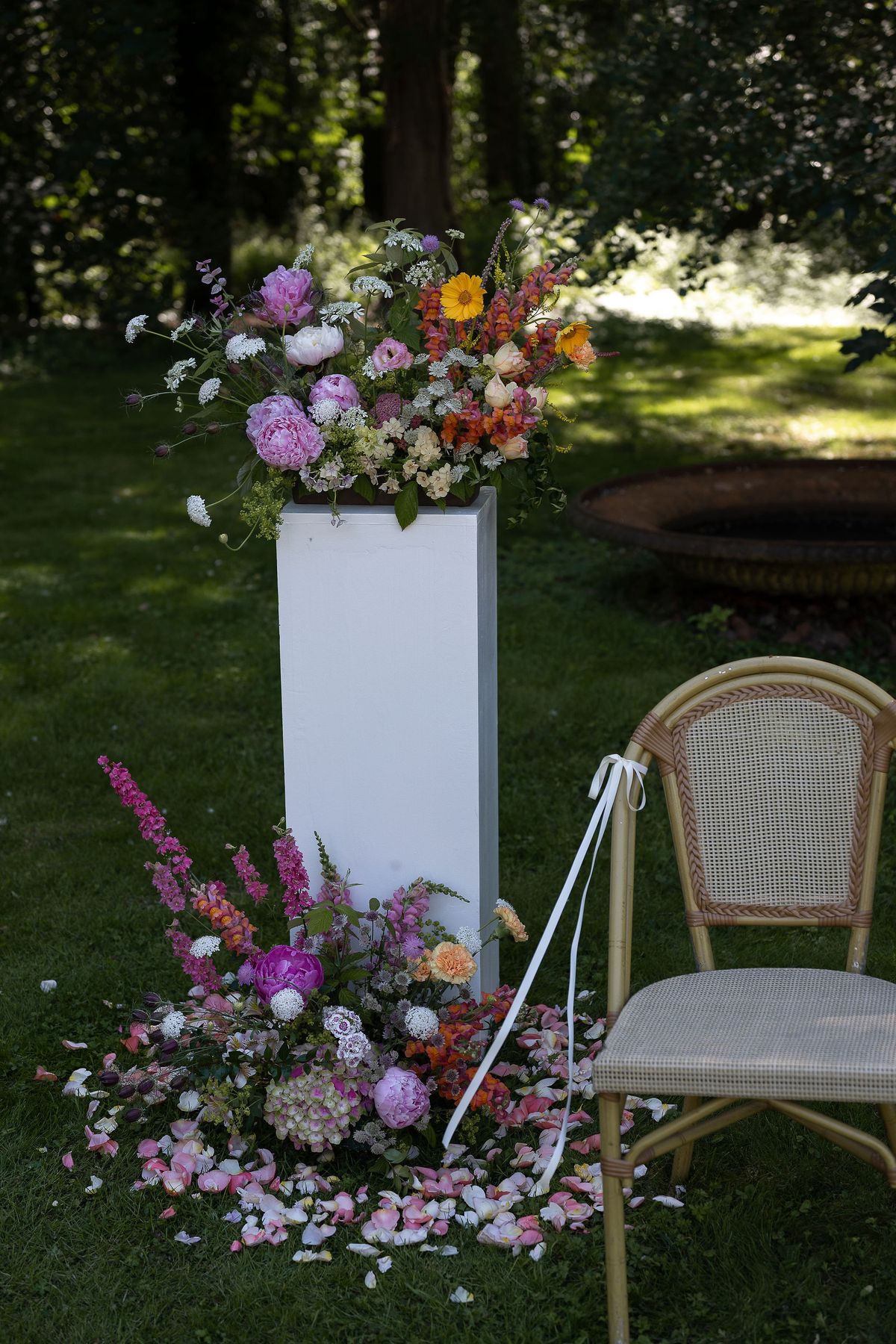A tall white plinth topped with colorful flowers stands on grass, with scattered petals at its base and a wicker chair beside it.