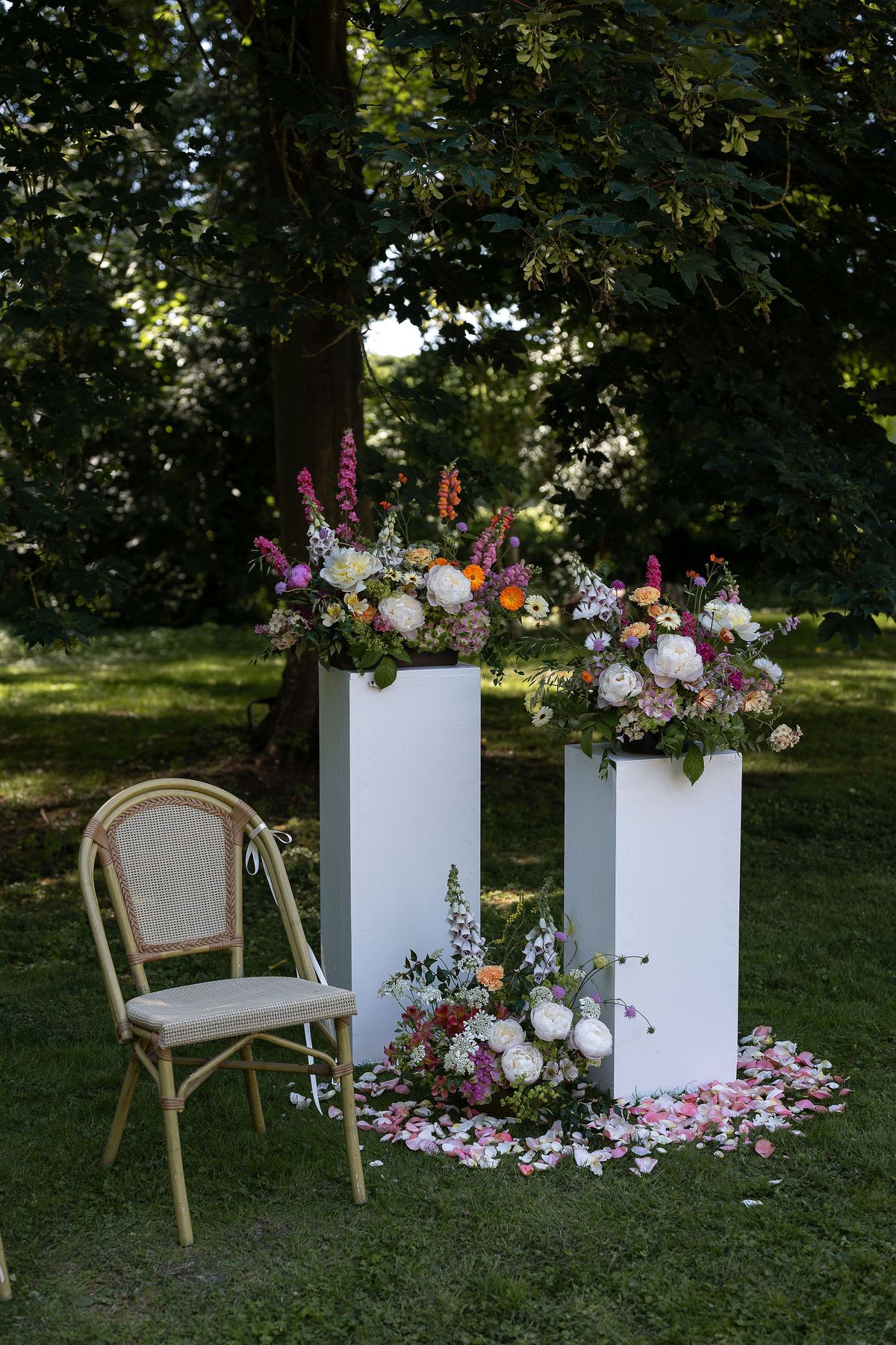 Two white pedestals topped with colorful flower arrangements stand on a lawn, with scattered petals at their base and a wooden cane chair beside them.