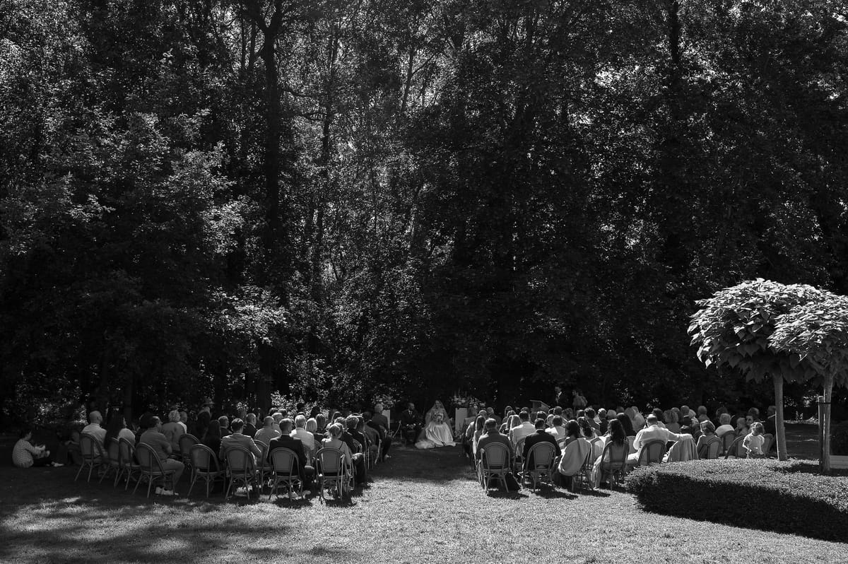 Guests seated on chairs in two groups on a lawn face a couple at the centre, beneath tall dark trees, in black and white.