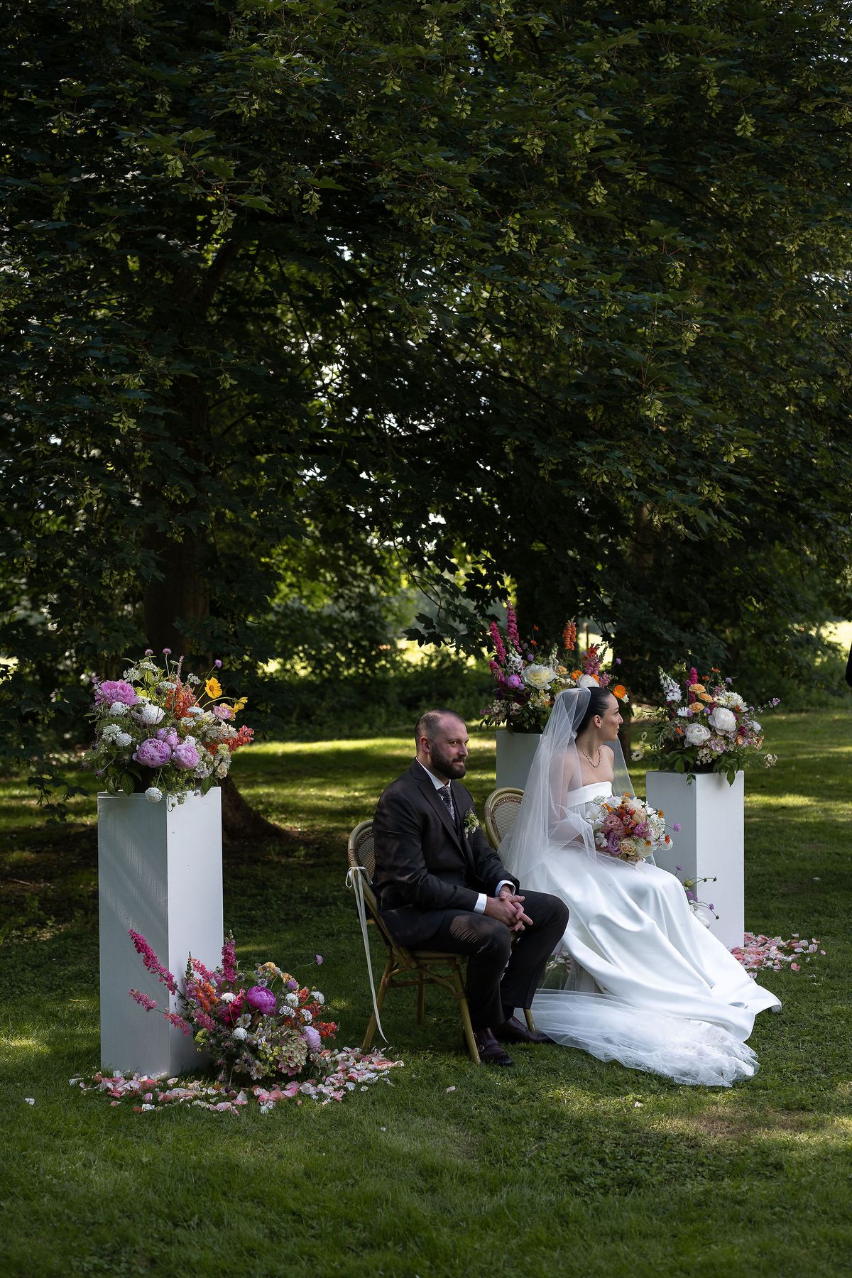 The groom and bride sit next to each other on chairs on the lawn, flanked by white plinths with flowers beneath large trees.