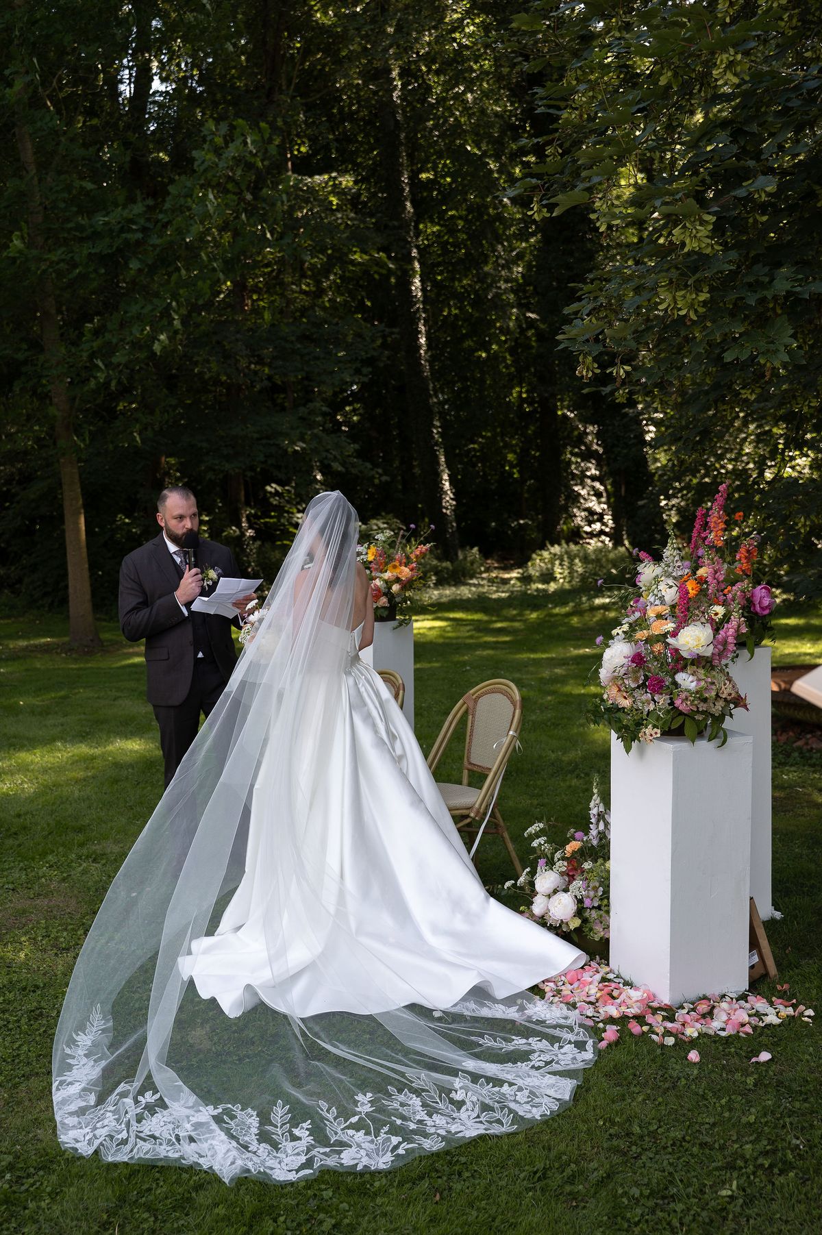 The bride in a wedding gown with a long lace-edged veil stands on the lawn facing an officiant, beside a tall flower-topped white plinth.