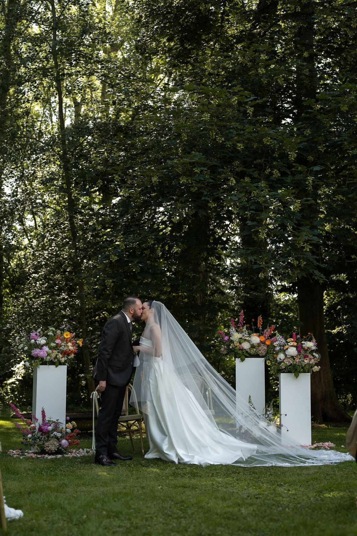 The couple kisses on the lawn under tall trees, the bride's long veil trailing behind, flanked by white plinths topped with flowers.