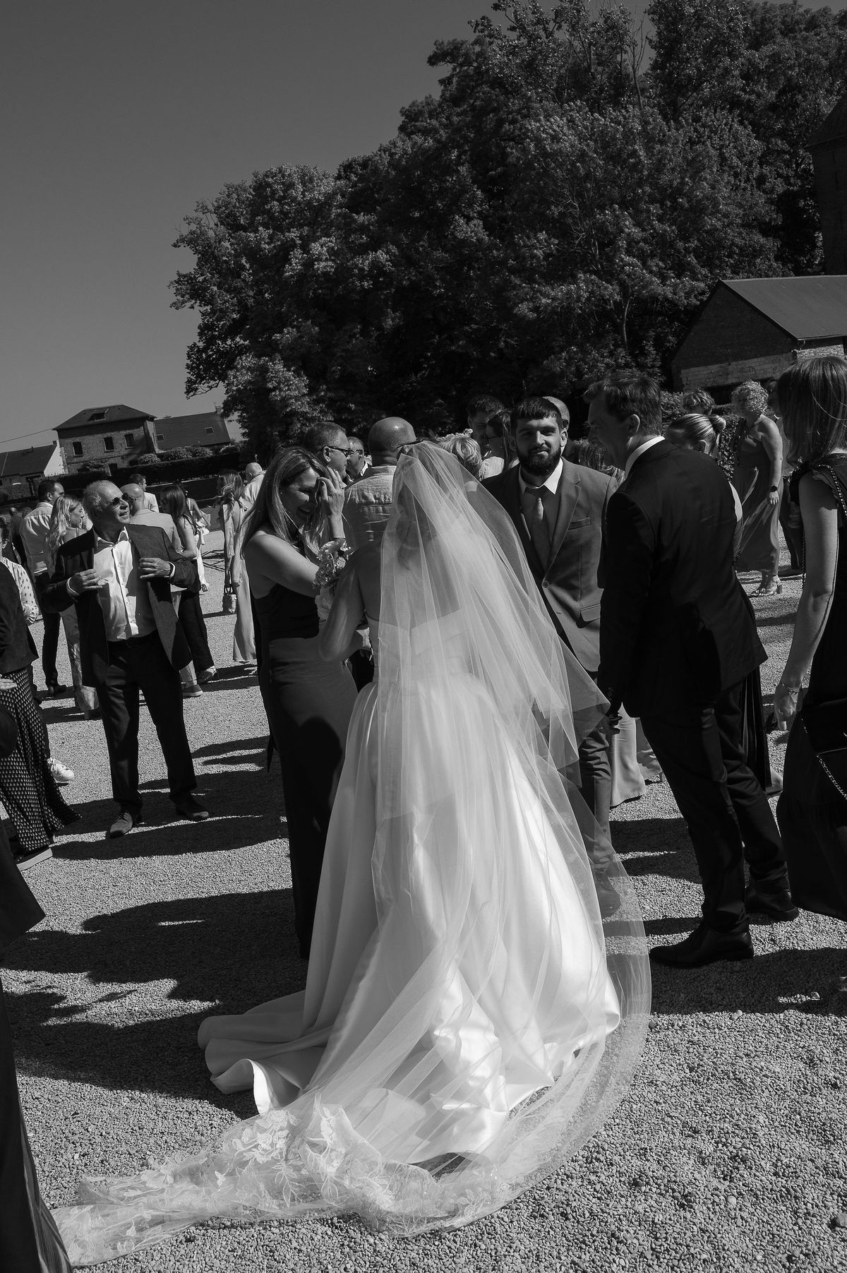 In black and white, the bride seen from behind in a long veil stands among guests chatting on a gravel courtyard.