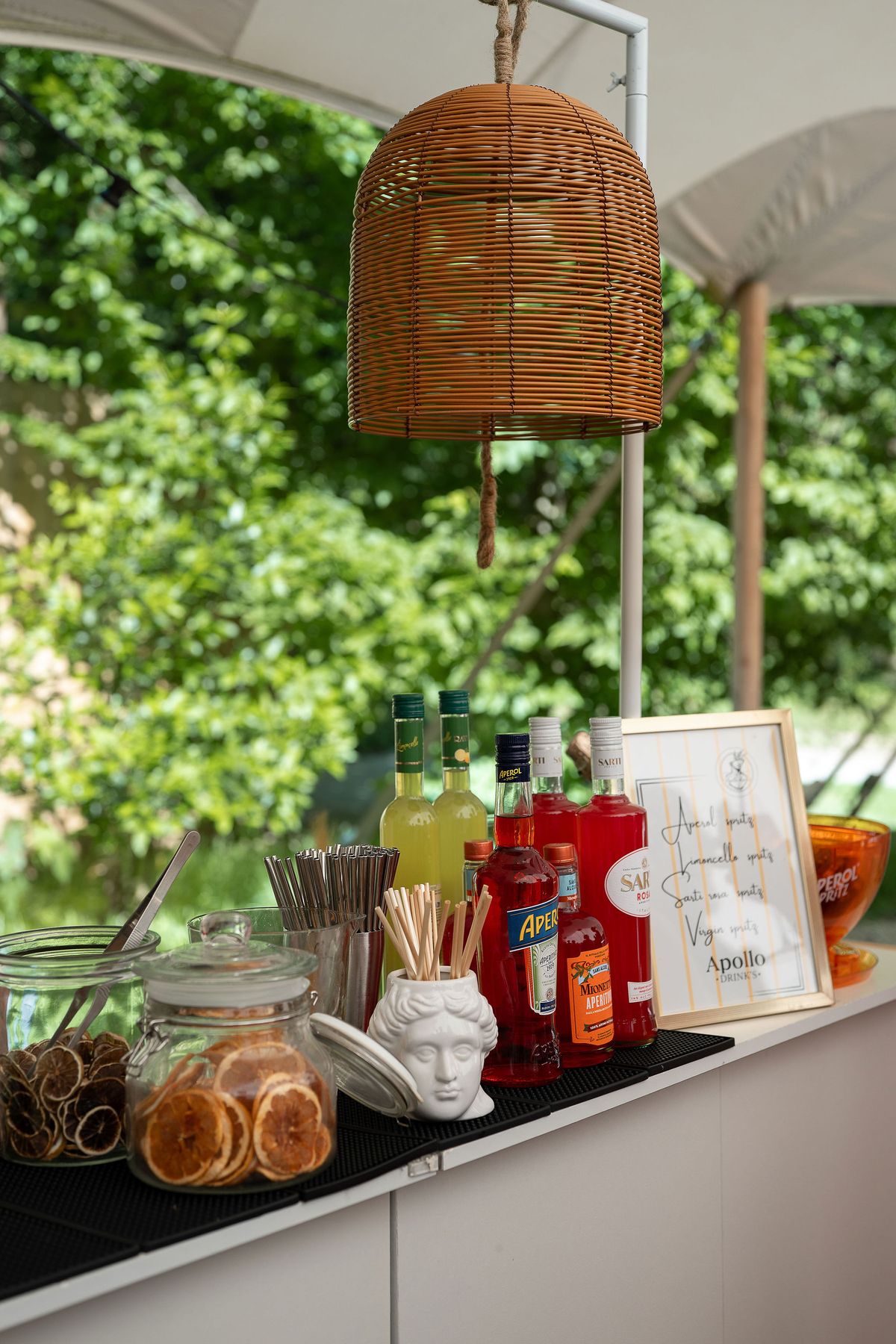 A bar counter under a tent holds liquor bottles, jars of dried orange slices, a framed menu sign and a hanging wicker lampshade.