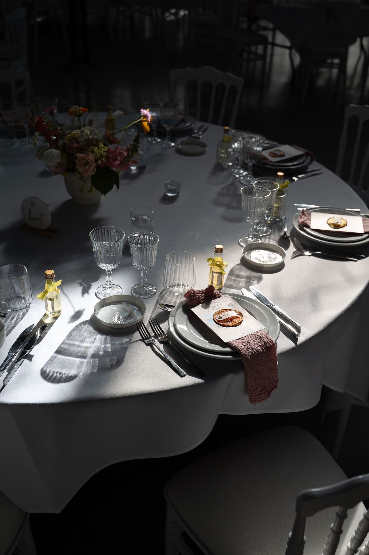 A round table with a white cloth is set with plates, glasses, a small flower arrangement and napkins in dim light.