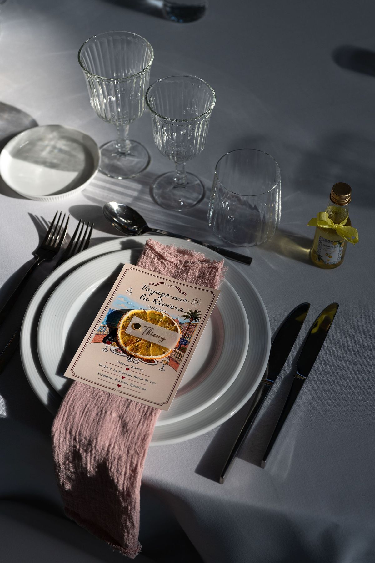 A white plate holds a menu card on a pink napkin, with cutlery and several glasses on a white tablecloth.