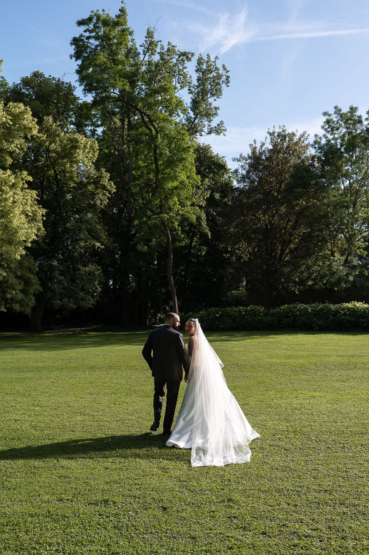 Seen from behind, the couple walks across a green lawn toward trees under a blue sky, the bride's veil trailing.