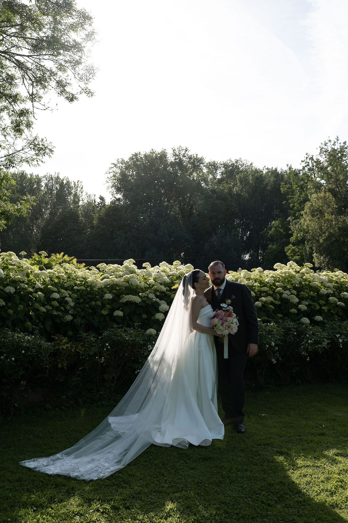 The bride in a white gown with a long veil embraces the groom on a lawn in front of a tall green hedge.