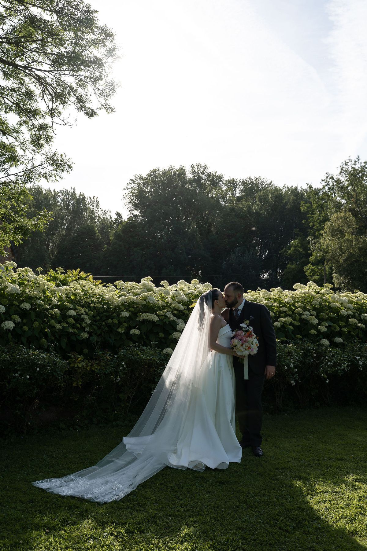 A bride in a long veil and a groom in a dark suit kiss on a lawn in front of a tall green hedge.