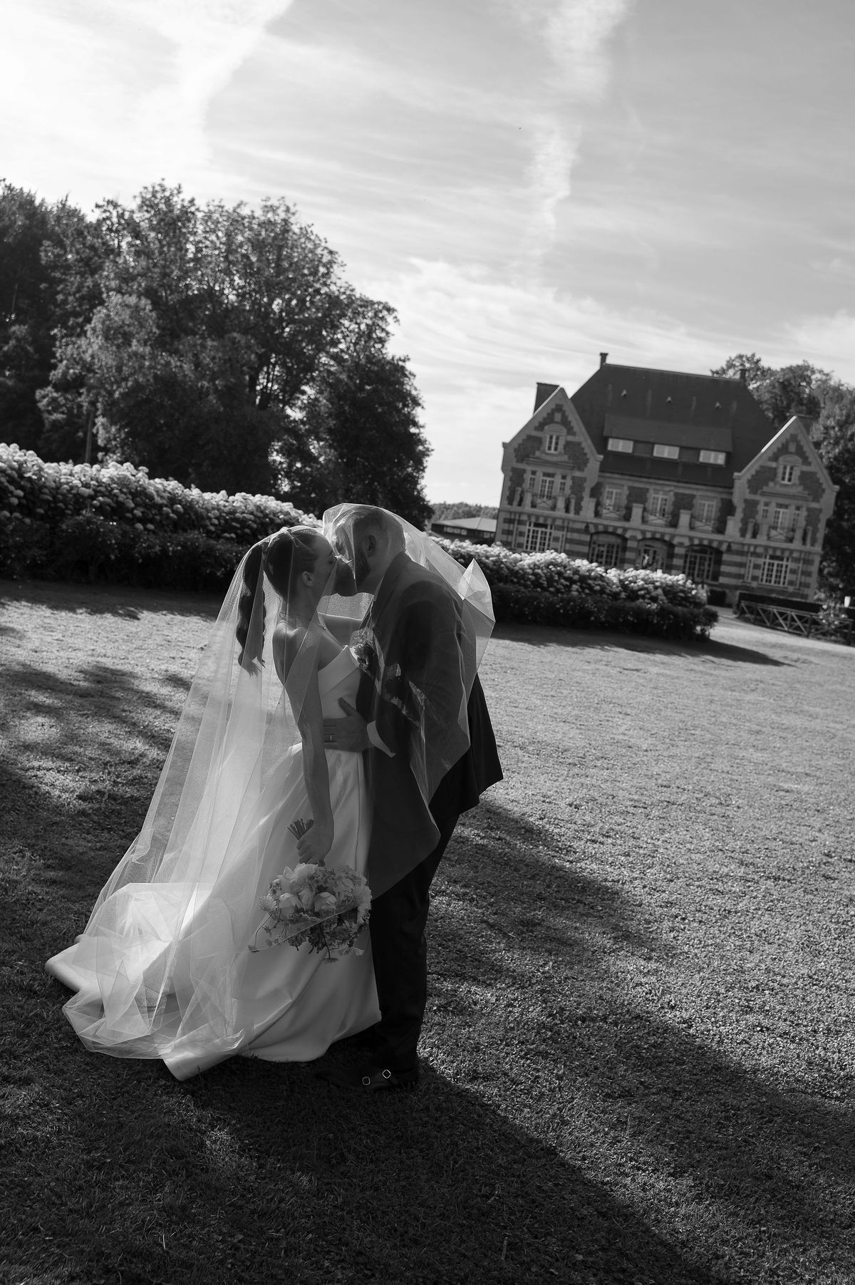 Black and white image of the couple leaning together on a lawn, the bride's veil flowing, with a chateau and trees behind.