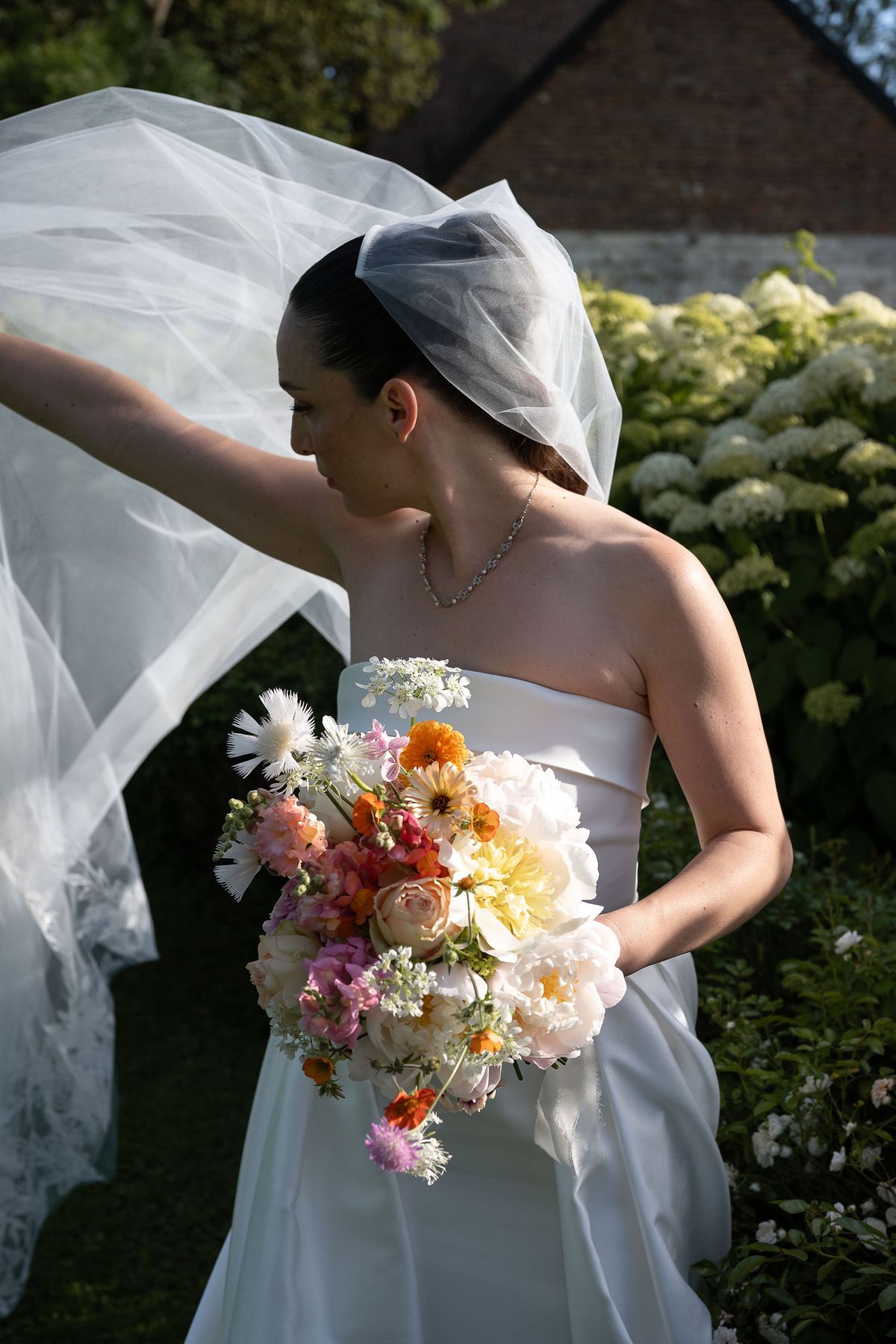 The bride lifts her flowing veil with one hand and holds a colorful bouquet in front of a flowering hedge.