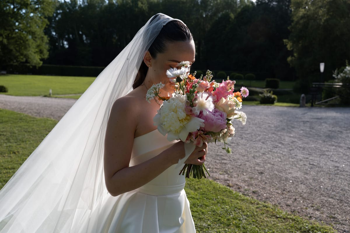 The bride in a strapless gown and long veil holds a pink and white bouquet to her face beside a gravel path.