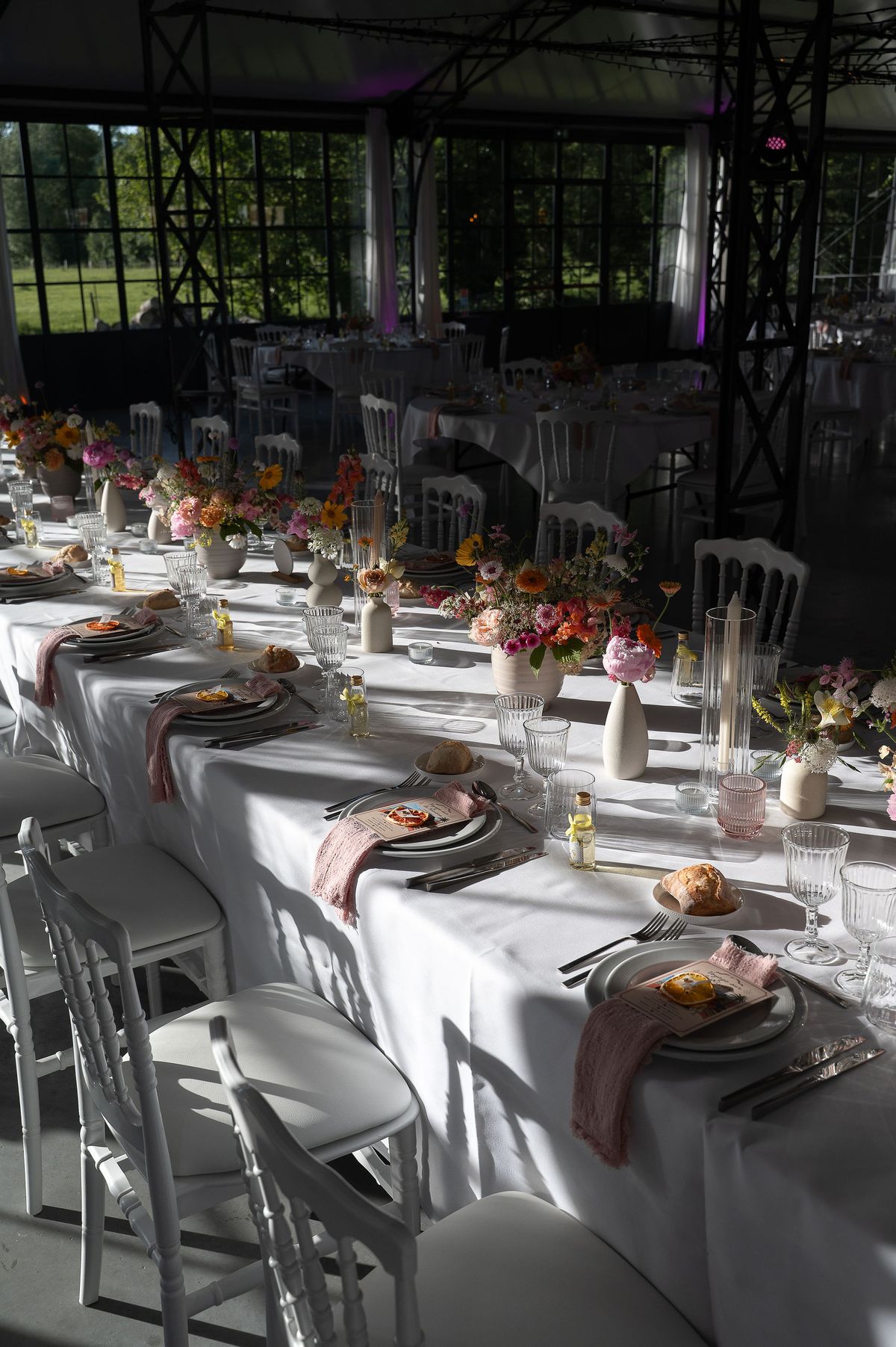 Long tables with white cloths, flower arrangements, plates and pink napkins stand in a bright hall with large windows.
