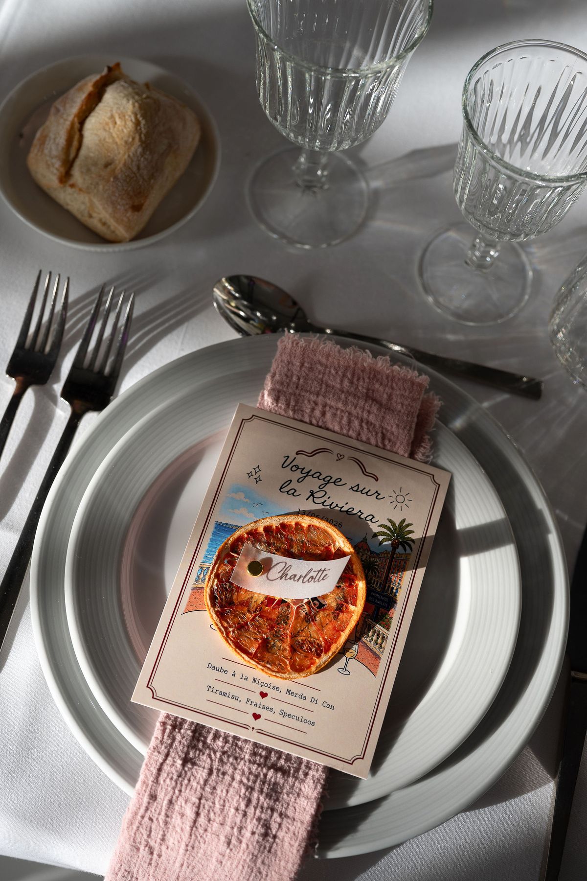 A plate with cutlery, a bread roll, glasses and a colorful illustrated menu card tucked in a pink napkin on a white tablecloth.