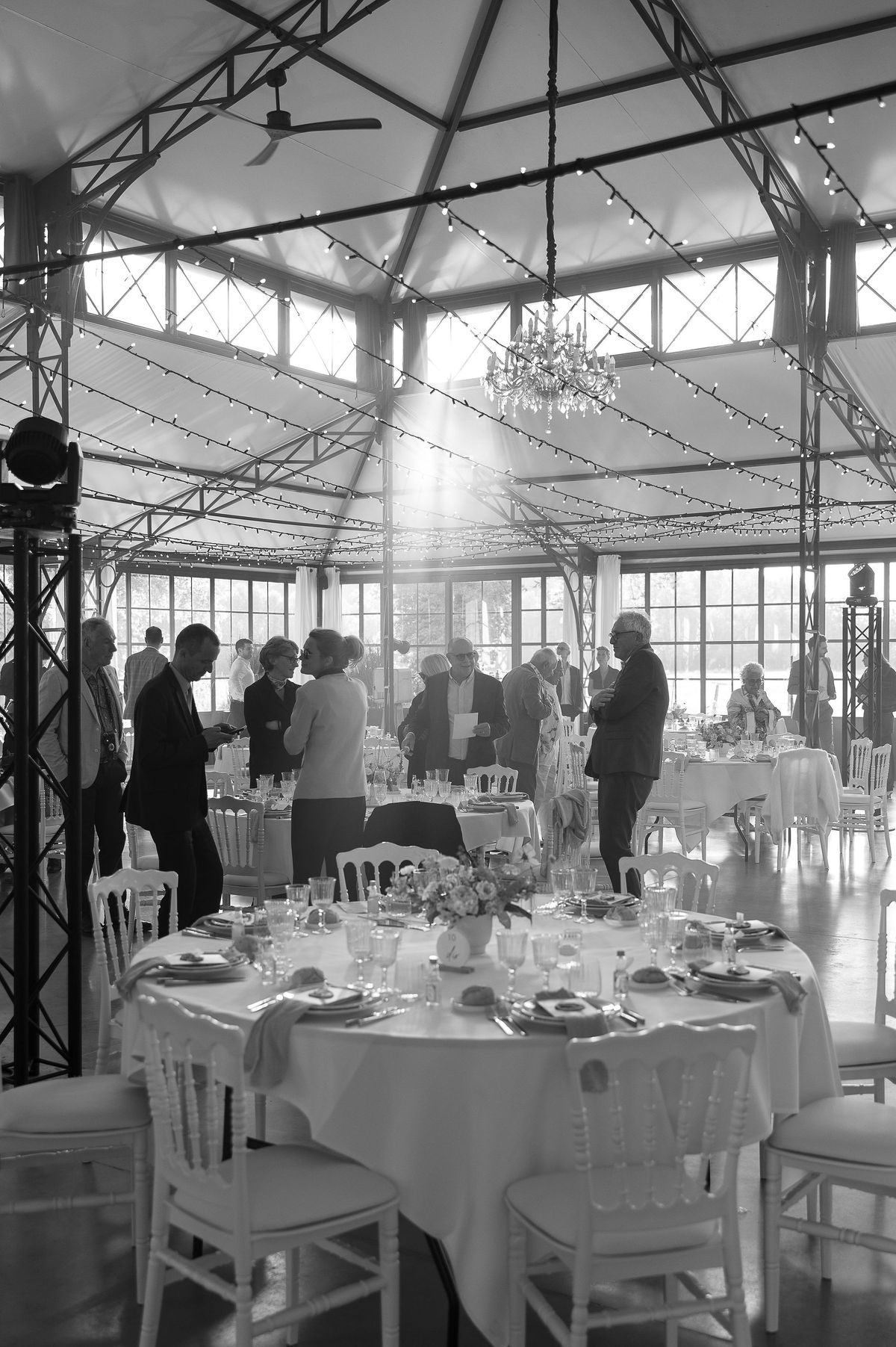 Guests stand talking among round tables laid with flowers and glasses in a bright glass-walled hall hung with fairy lights.
