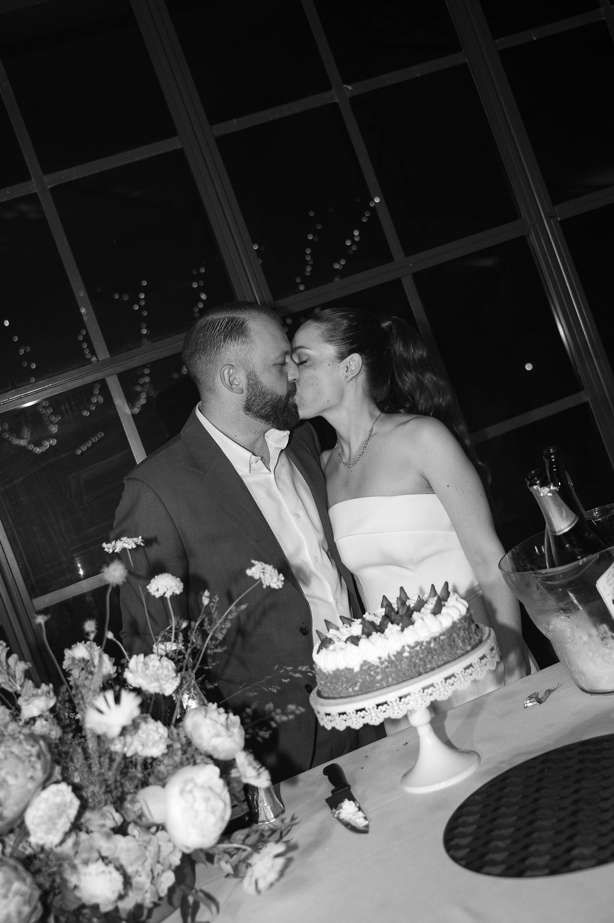 Black and white image of the couple kissing over a cake on a table with flowers in the foreground, in a glass-walled room.