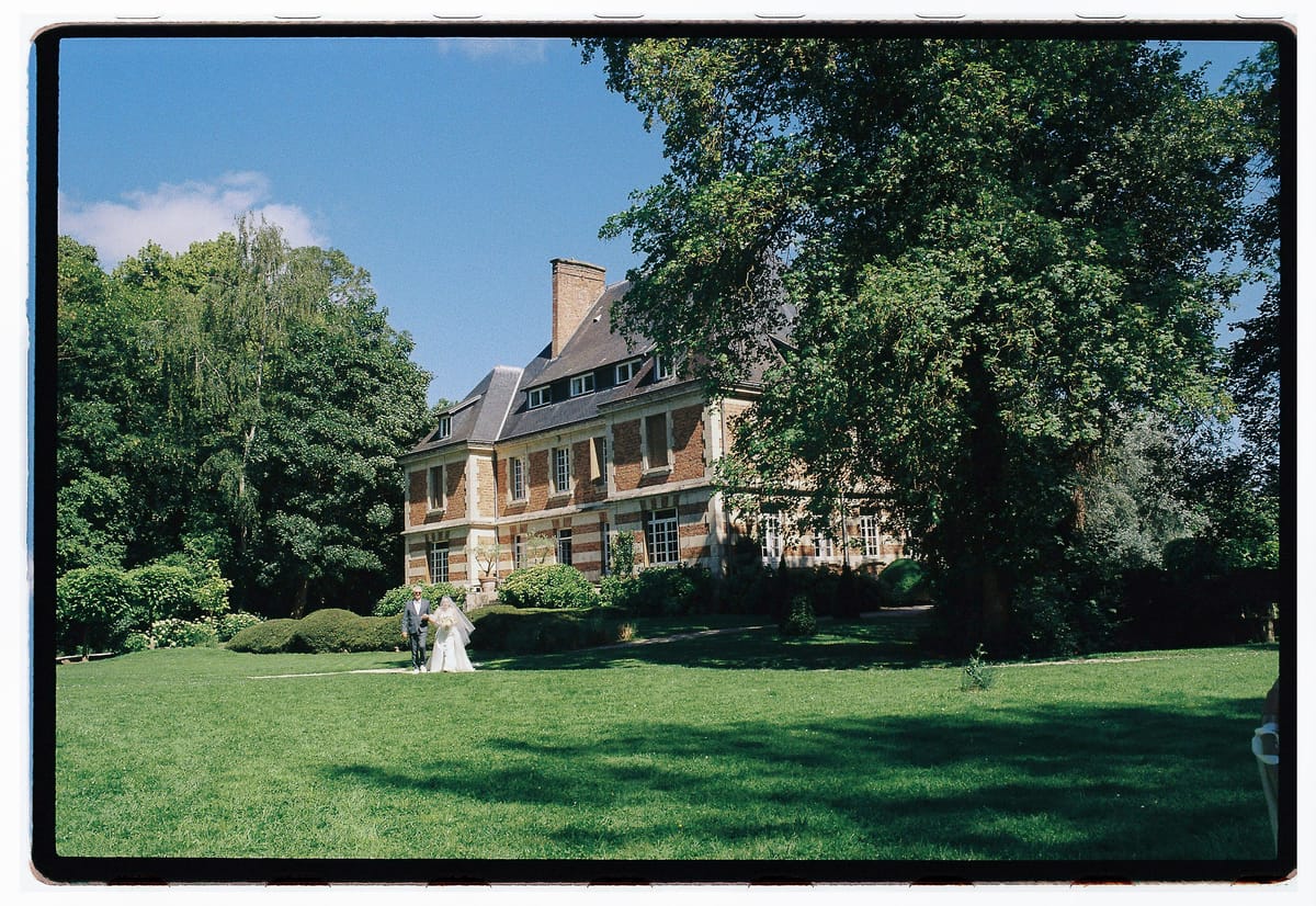 A large brick chateau stands behind a wide lawn and a big tree, with two tiny figures in white near the building.