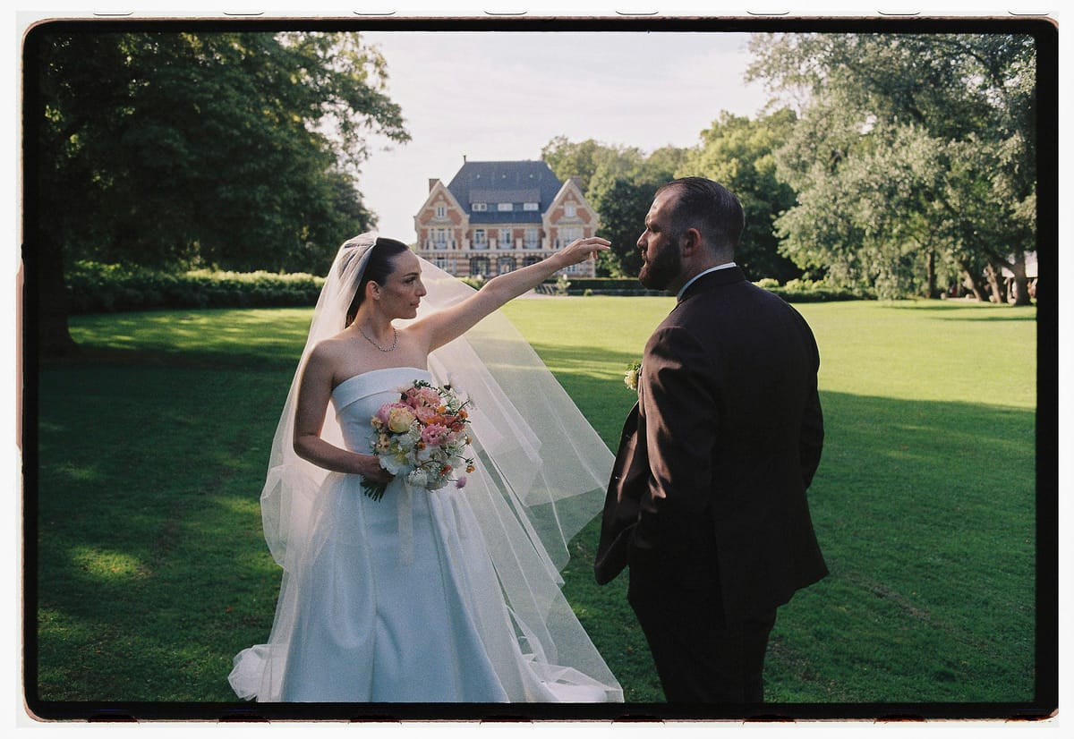 The bride, holding a bouquet, points across the lawn toward a distant chateau while the groom in a dark suit faces her.