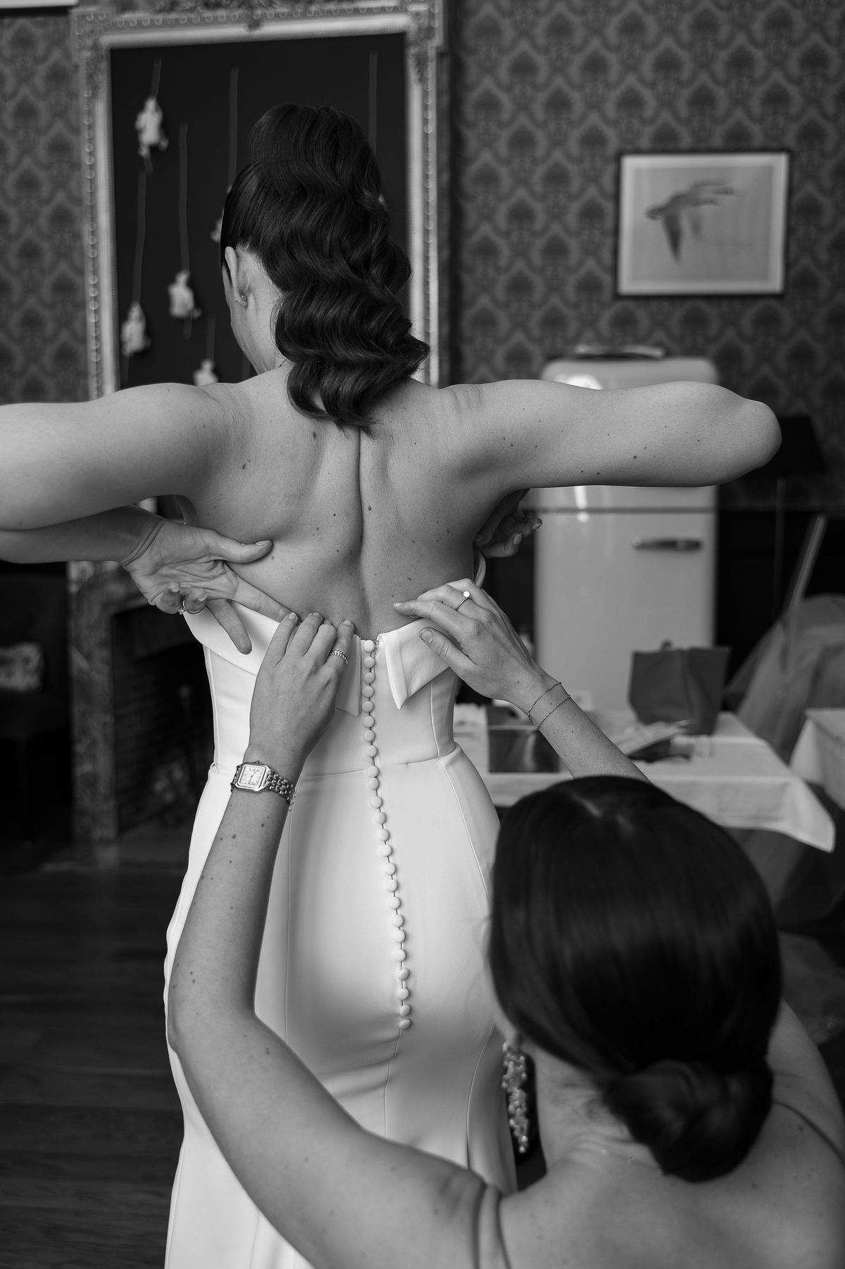 Black and white view from behind of the bride in a buttoned gown while another woman fastens it in a patterned-wallpaper room.