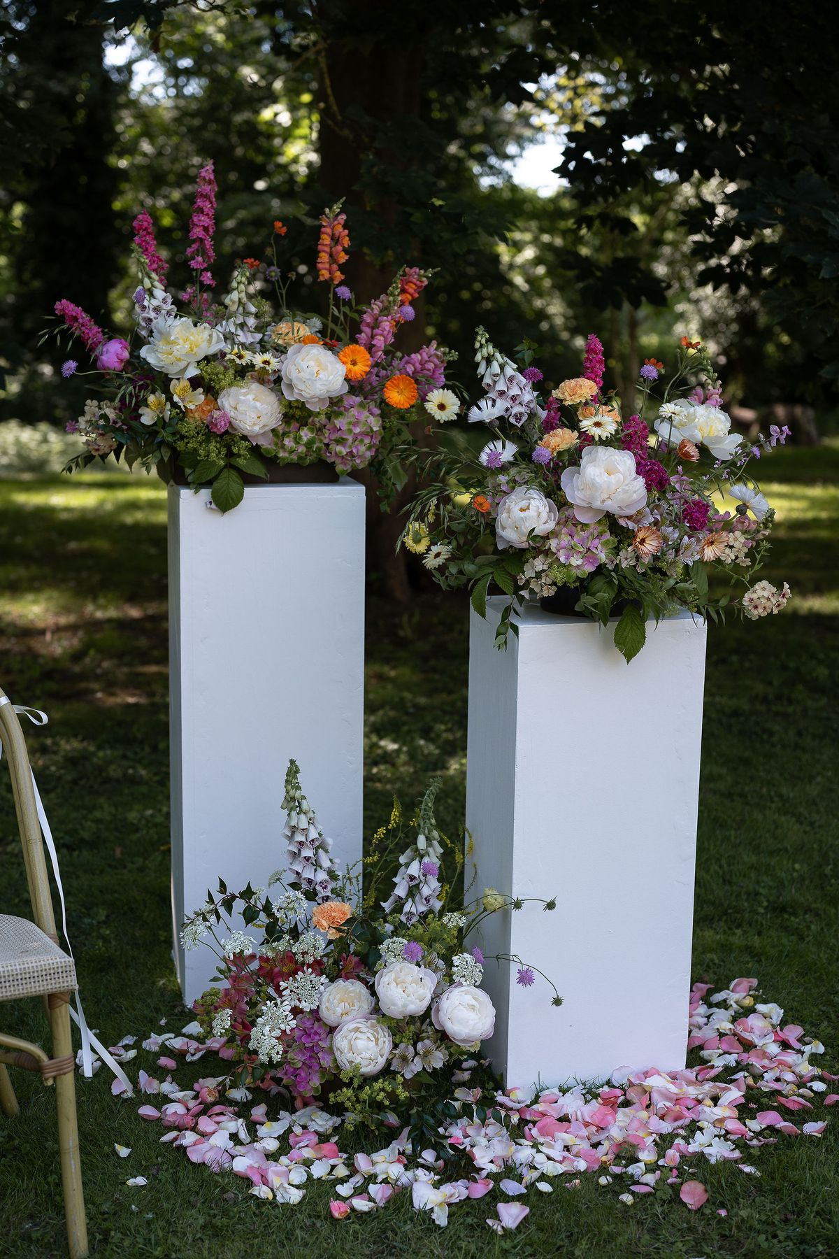 Two white plinths hold lush bouquets of pink, orange and white flowers, with petals scattered on the grass beneath trees.