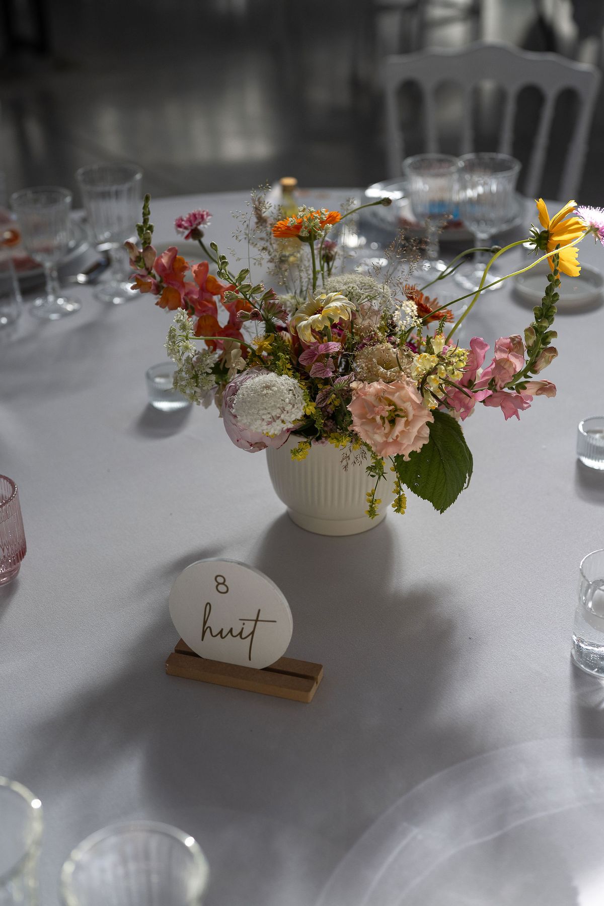 A small vase of pink, orange and white flowers on a round table with glasses and a handwritten table number card.
