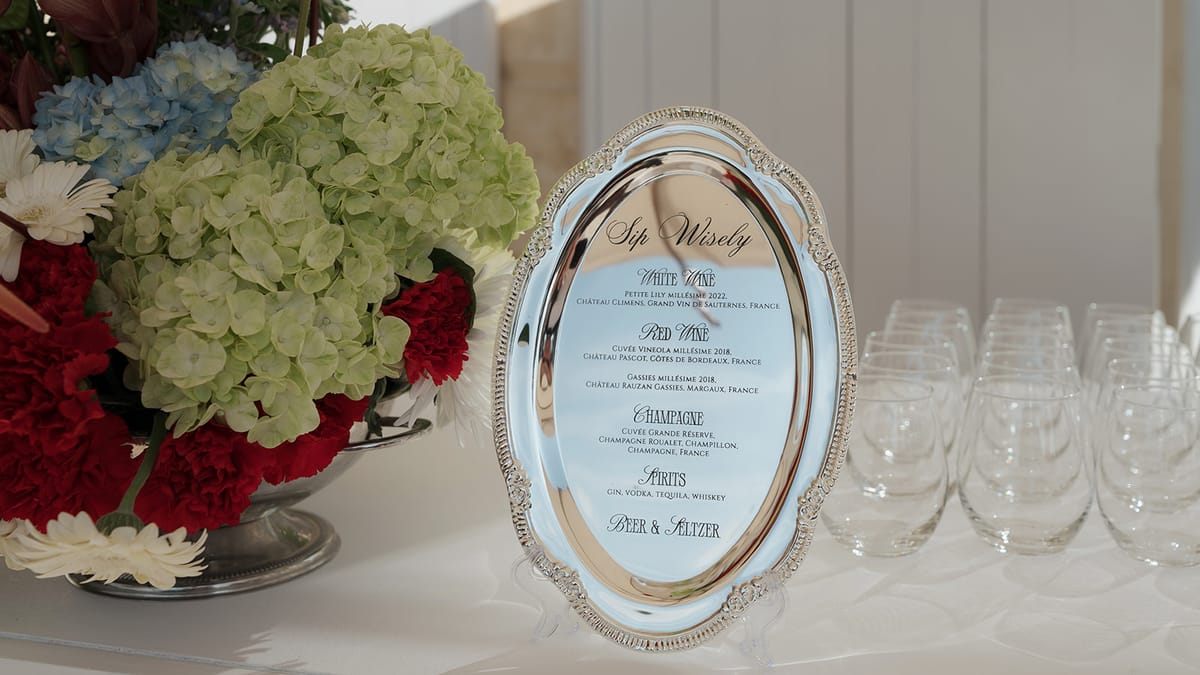 A "Sip Wisely" drinks list printed on an oval silver tray beside hydrangeas and red carnations
