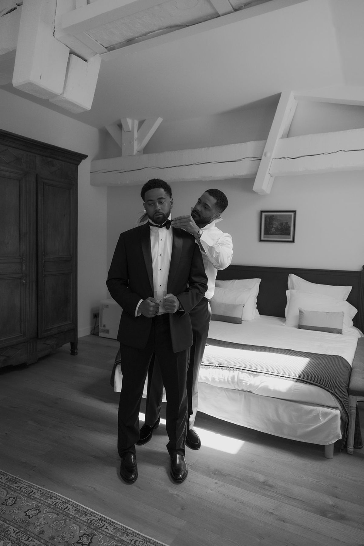 A groomsman helping Brandon with his collar in a bedroom, black and white