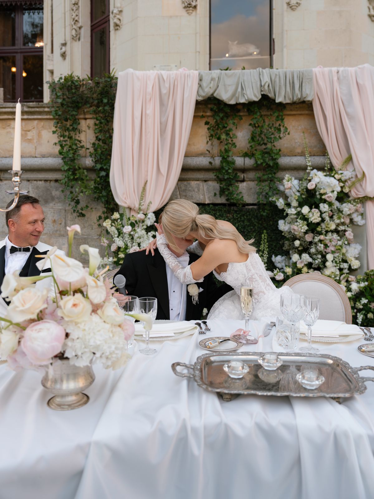 Kira hugs Steve at the top table beneath blush drapes and white florals at Chateau Challain