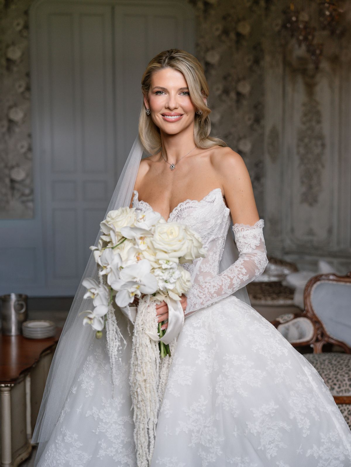 Kira with her orchid bouquet and lace gloves in a carved salon at Chateau Challain