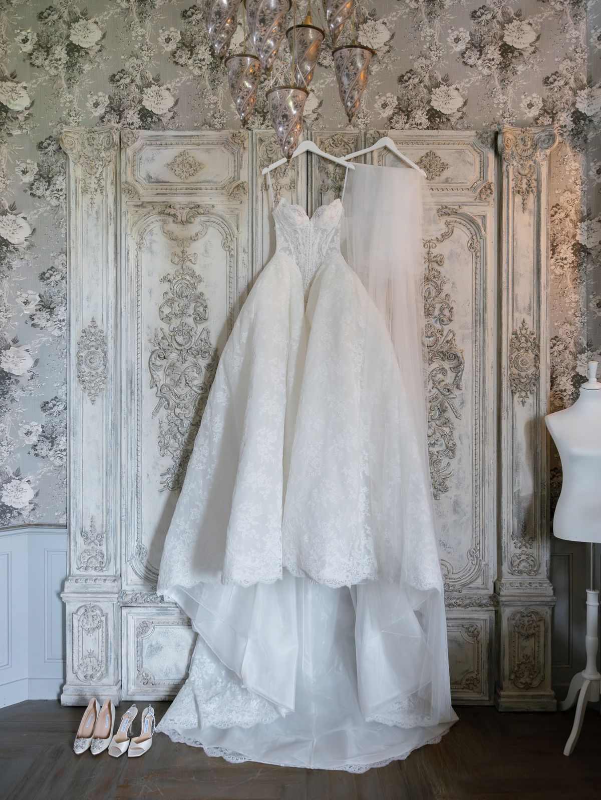 Kira's lace wedding gown hangs against carved panelling with bridal heels below at Chateau Challain