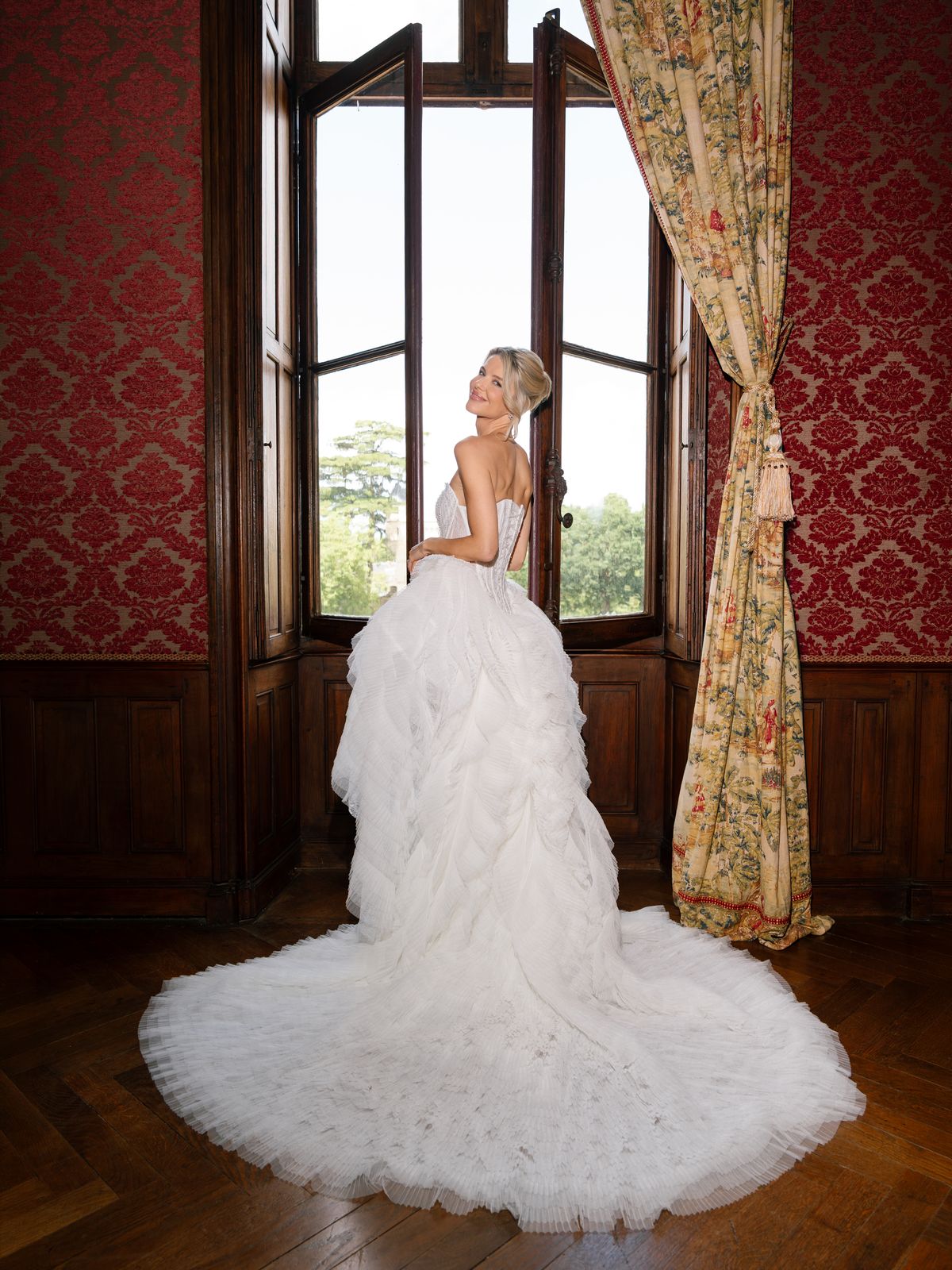 Kira at an open window in a red damask room at Chateau Challain, her tulle train spread behind her