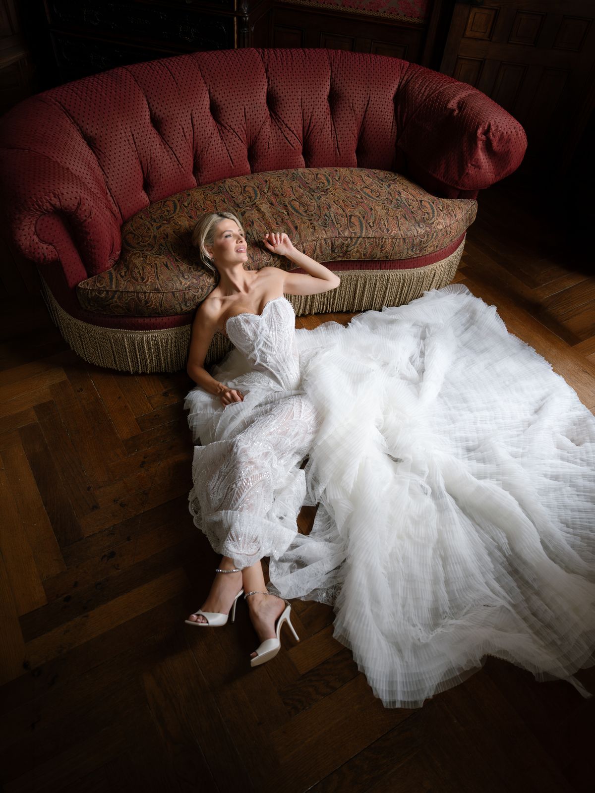 Kira reclines against a red velvet sofa with her tulle train spread across the parquet at Chateau Challain