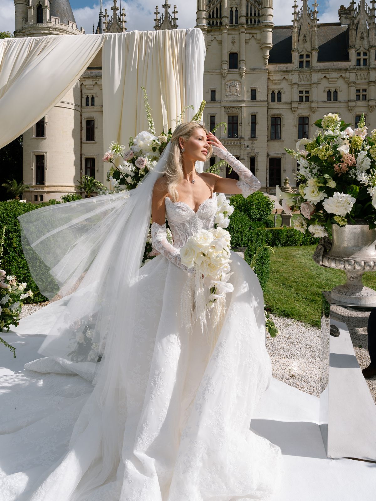 Kira in a lace-gloved ball gown and long veil before the floral ceremony arch at Chateau Challain
