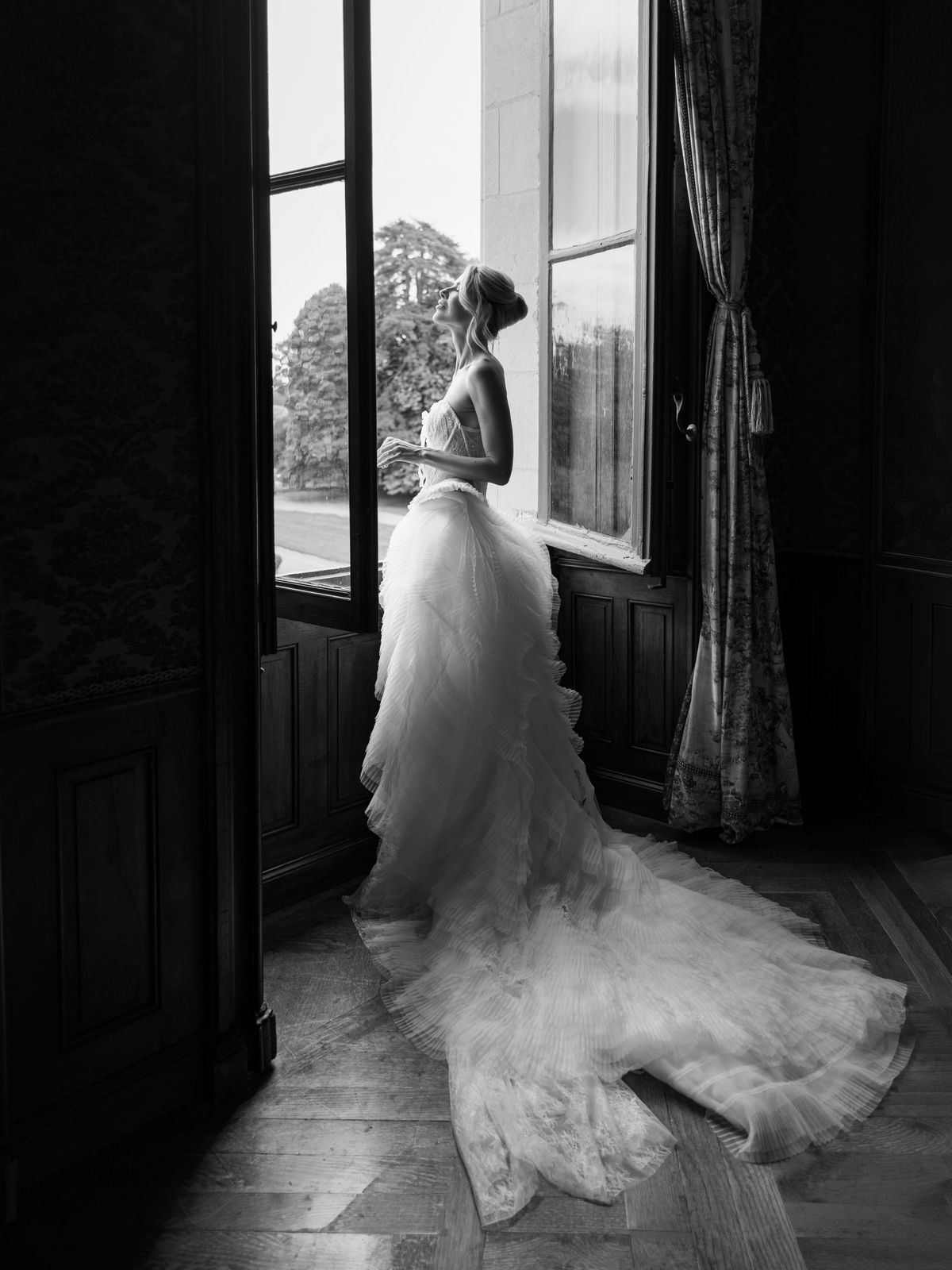 Kira stands at a tall window at Chateau Challain with her tulle train pooled on the floor