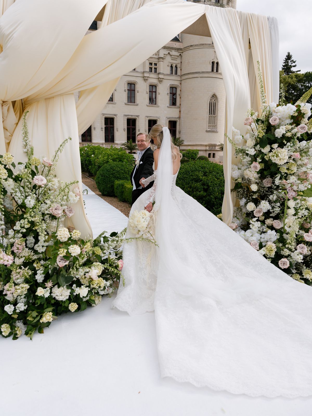 Kira reaches Steve at the draped floral ceremony arch in front of Chateau Challain