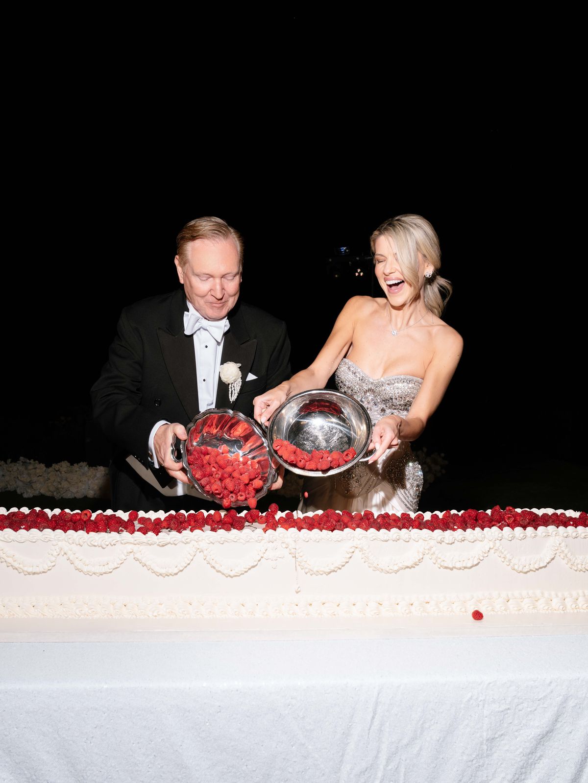 Kira and Steve laugh as they pour raspberries onto the wedding cake at night