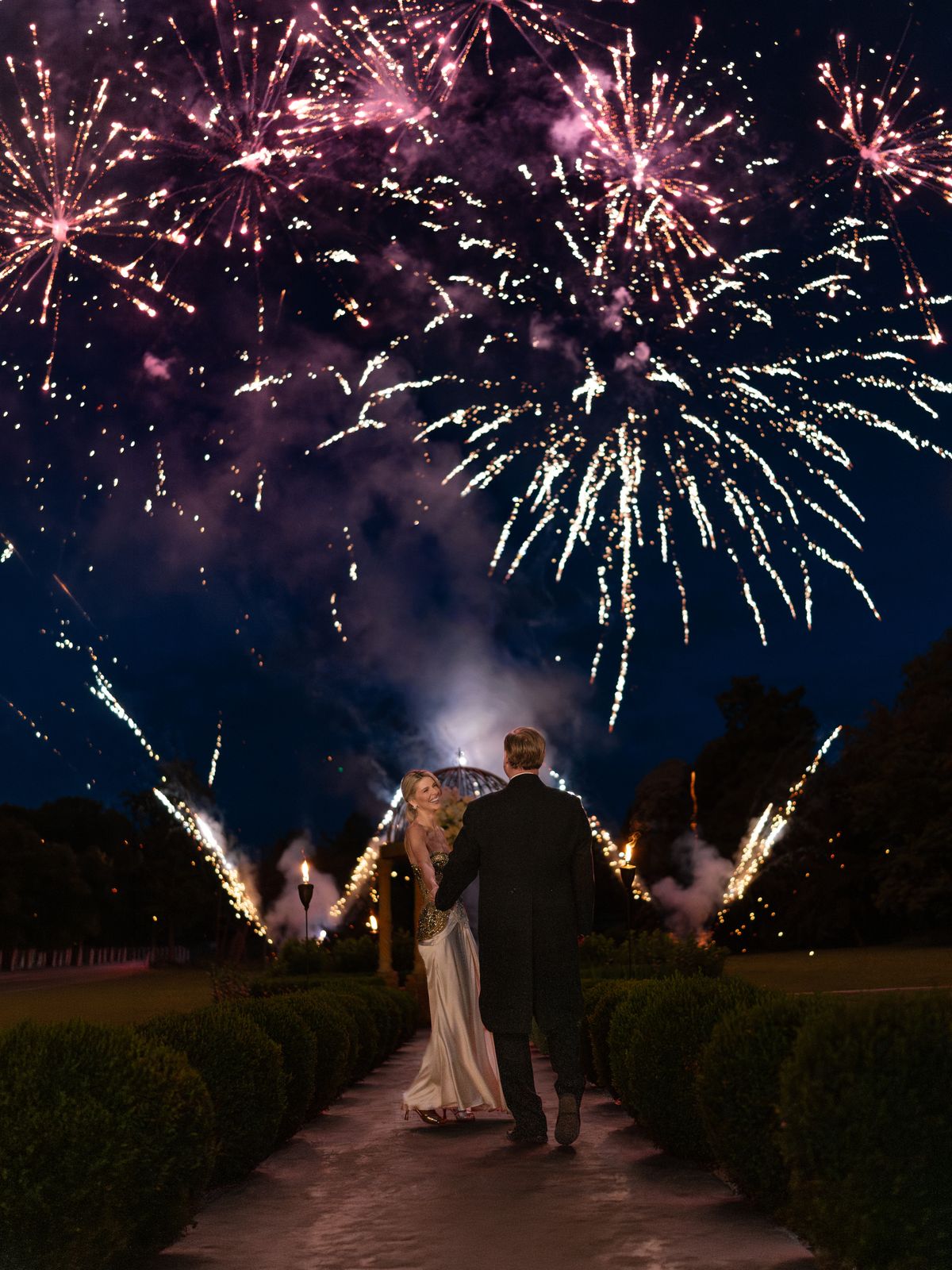 Kira and Steve walk down a lit garden path as fireworks burst above Chateau Challain
