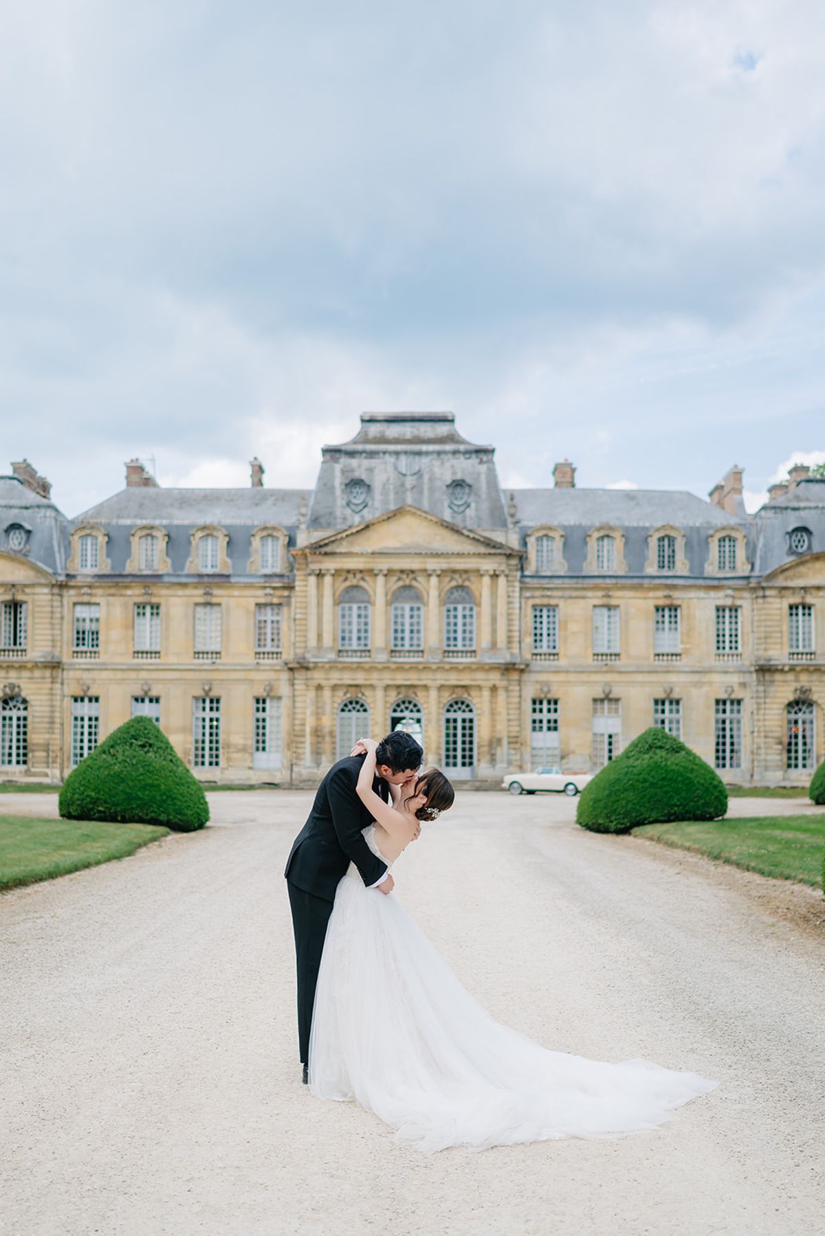 Liana and Ruiji embracing beside a vintage car with Château de Champlâtreux behind them