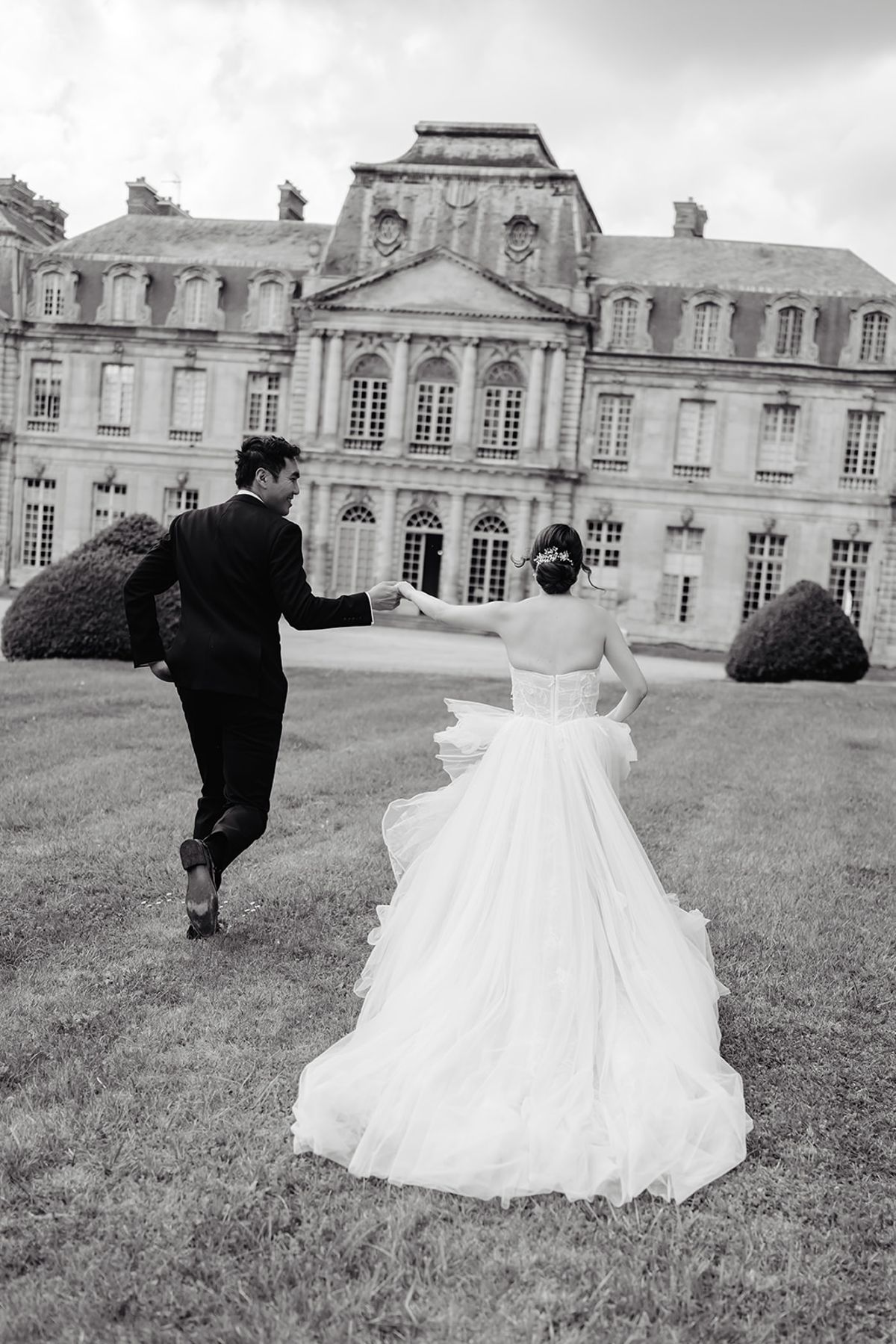 Liana and Ruiji running hand in hand toward Château de Champlâtreux, black and white