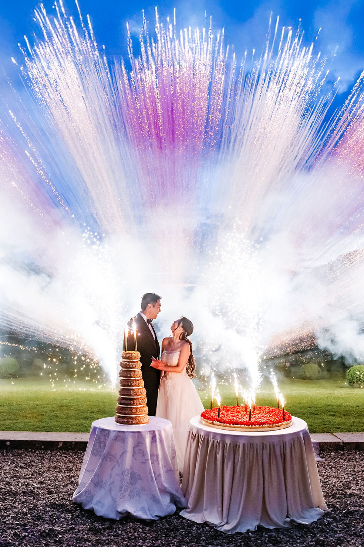 Liana and Ruiji with their cake as fireworks light up the night sky