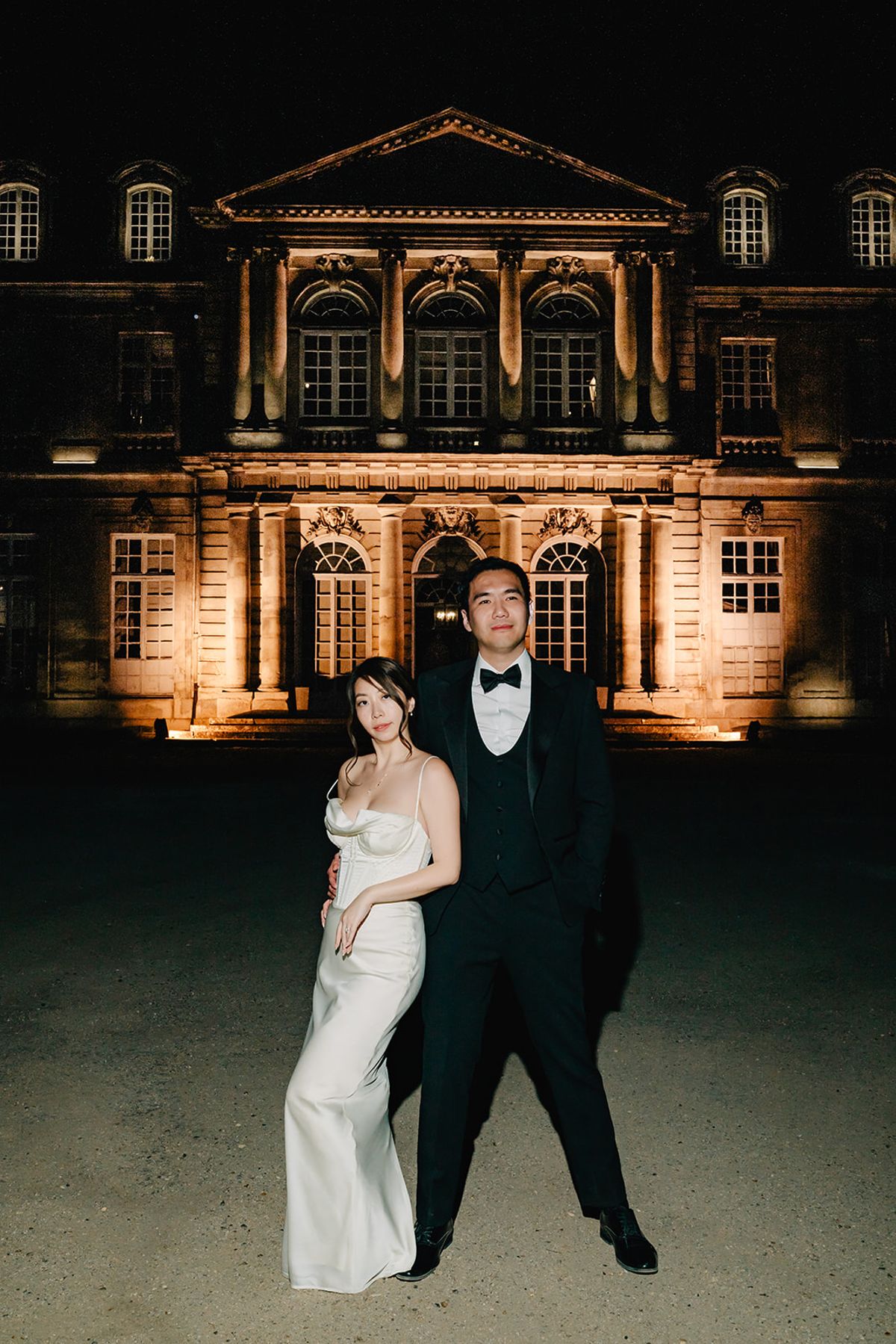 Liana and Ruiji posed together at night in front of the illuminated Château de Champlâtreux