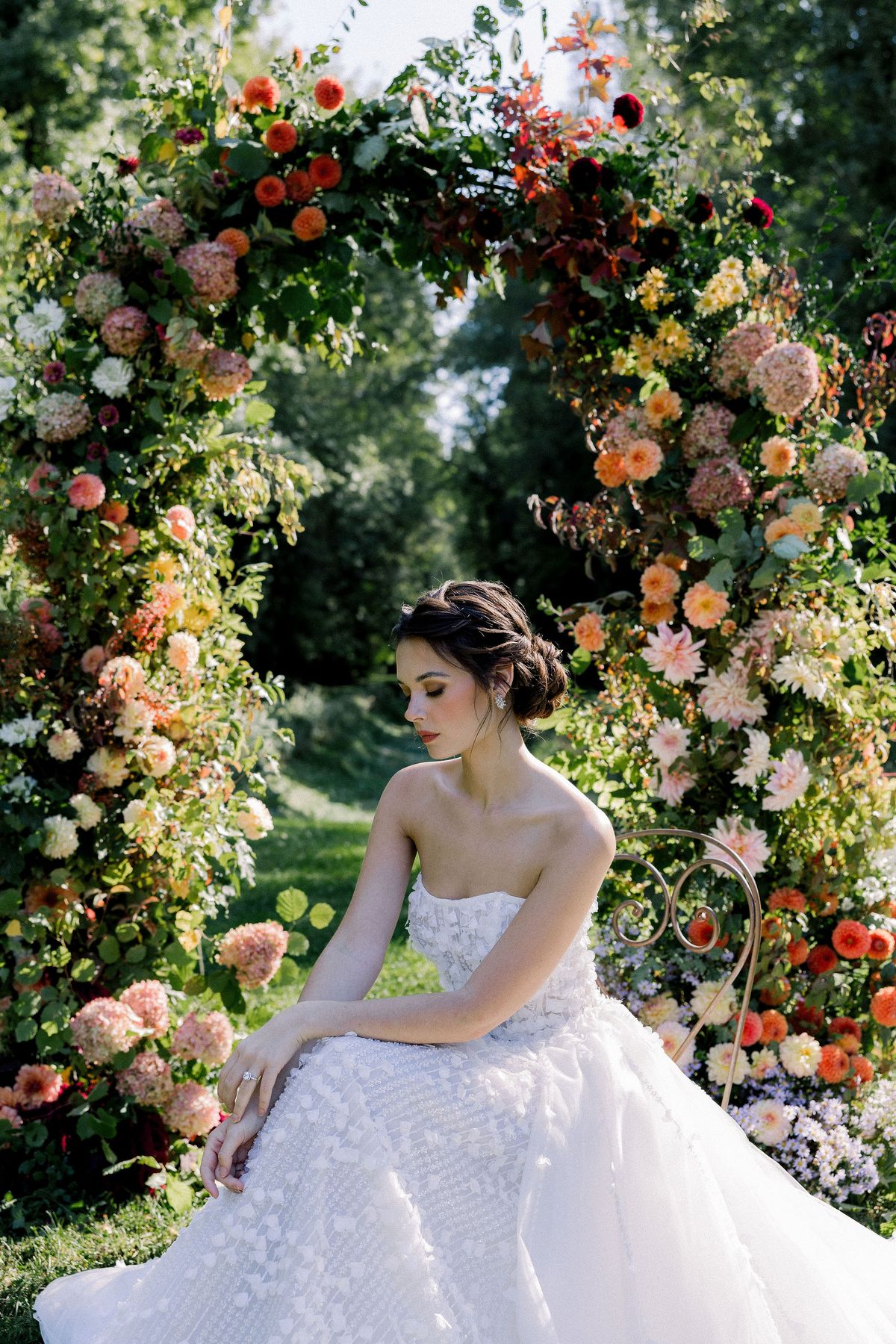 The bride in a strapless beaded gown sits with her hand on her knee in front of an arch of orange and pink flowers.