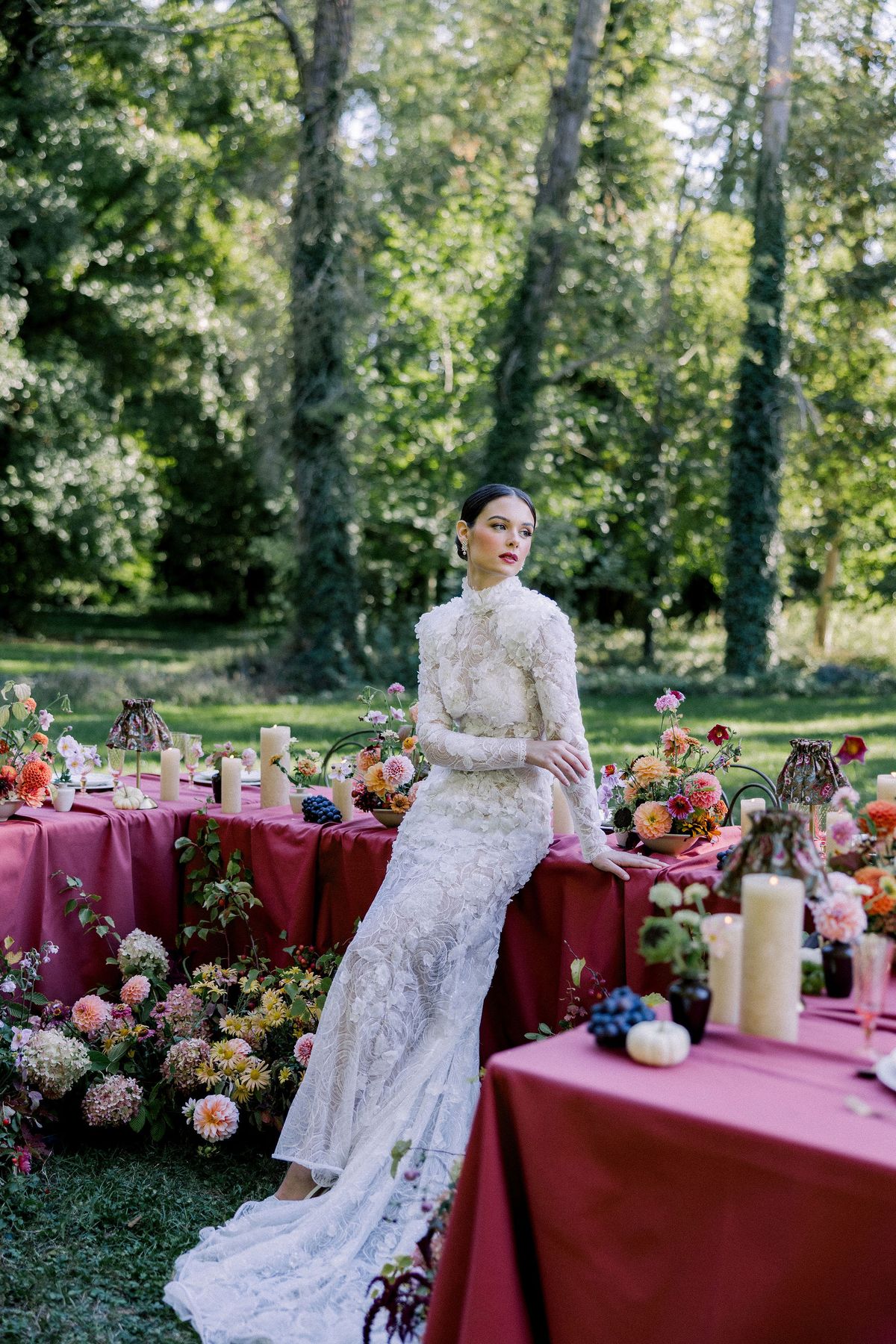 The bride in a lace gown leans on the edge of a burgundy table surrounded by flowers and candles.