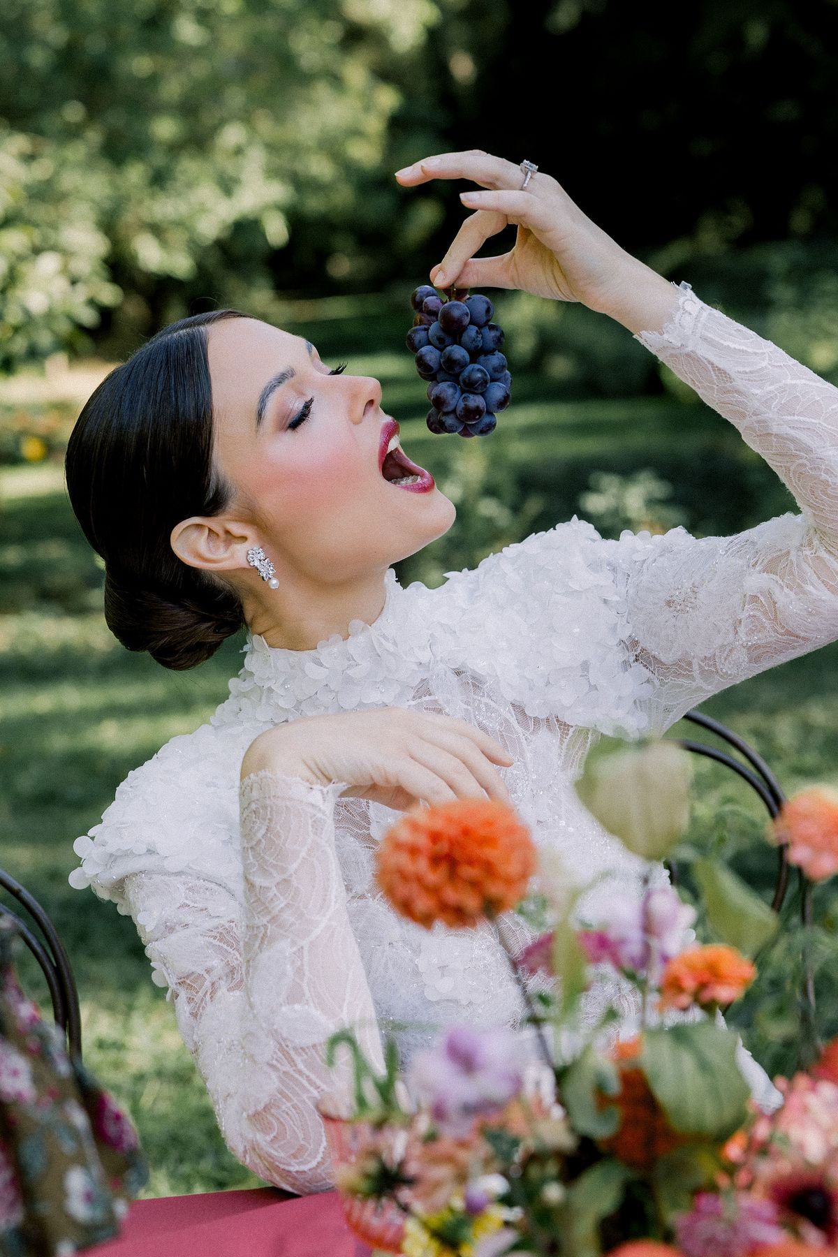 A bride in a lace gown tilts her head back and holds a bunch of dark grapes above her open mouth beside flowers.