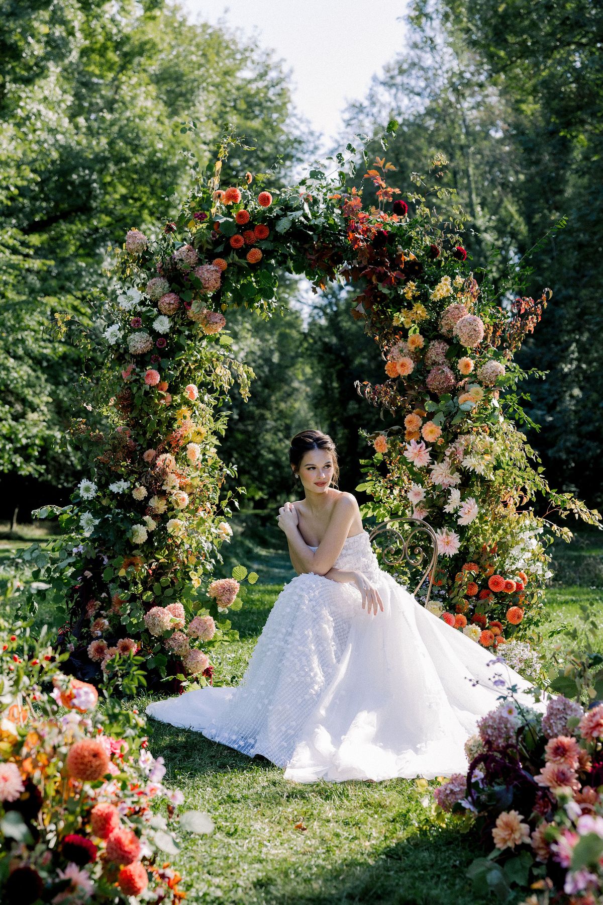 A bride in a strapless tulle gown sits on an iron chair beneath an arch of orange, pink and white flowers in a wooded garden.