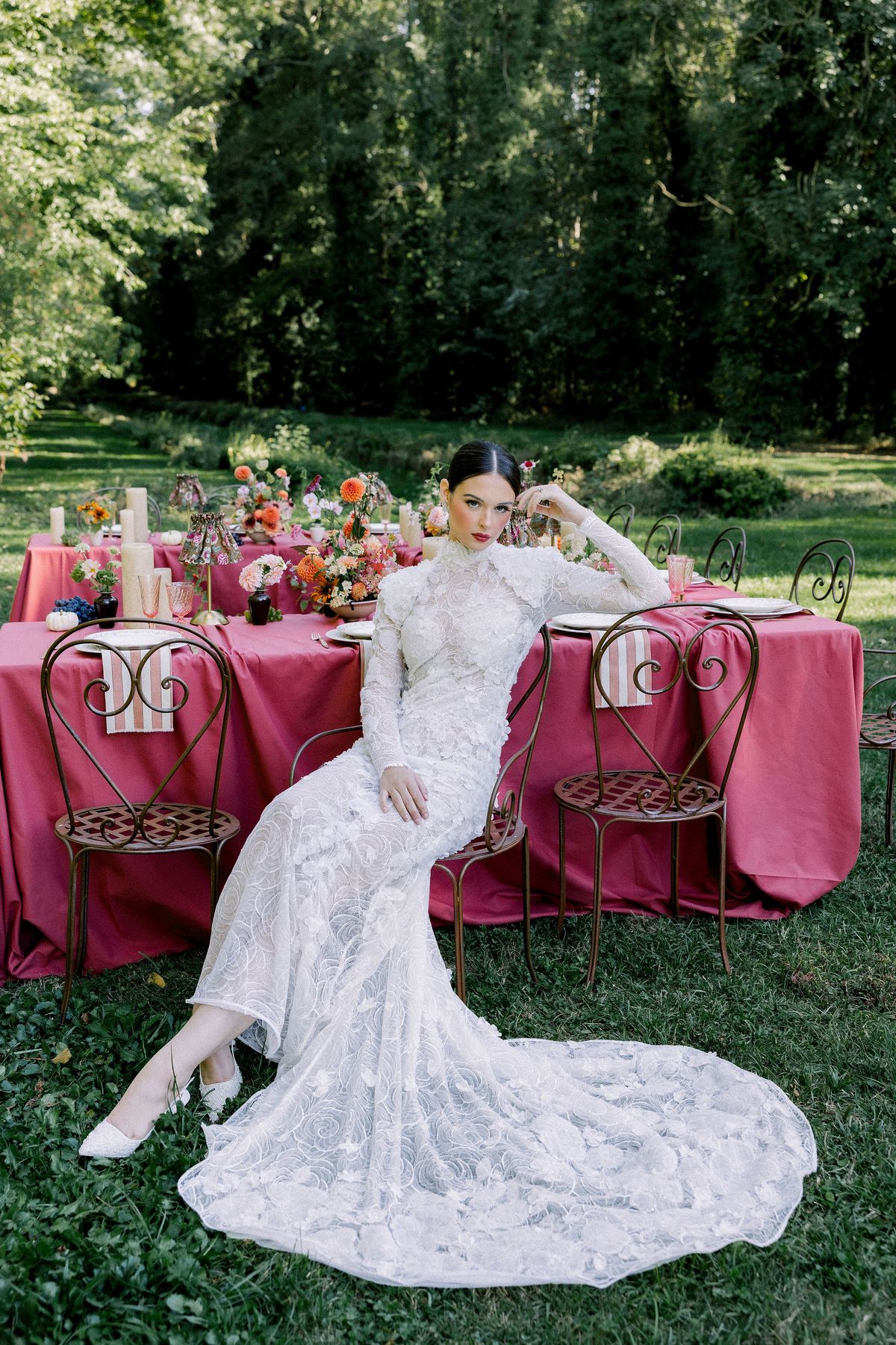 A bride in a long lace gown sits sideways on a chair at a flower-decked table with a raspberry cloth outdoors.