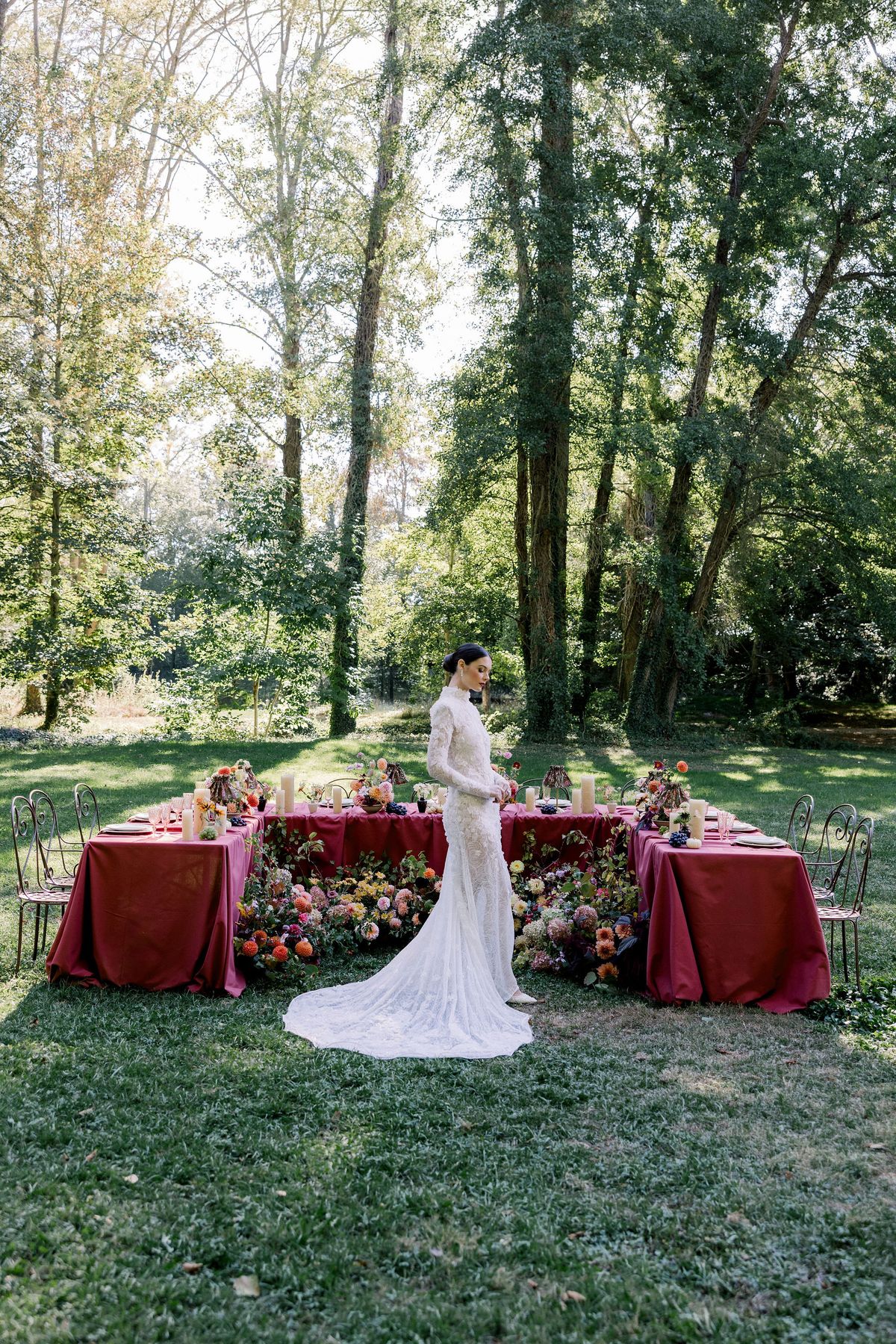 The bride in a lace gown with a train stands on the lawn in front of a burgundy table, with tall trees behind.