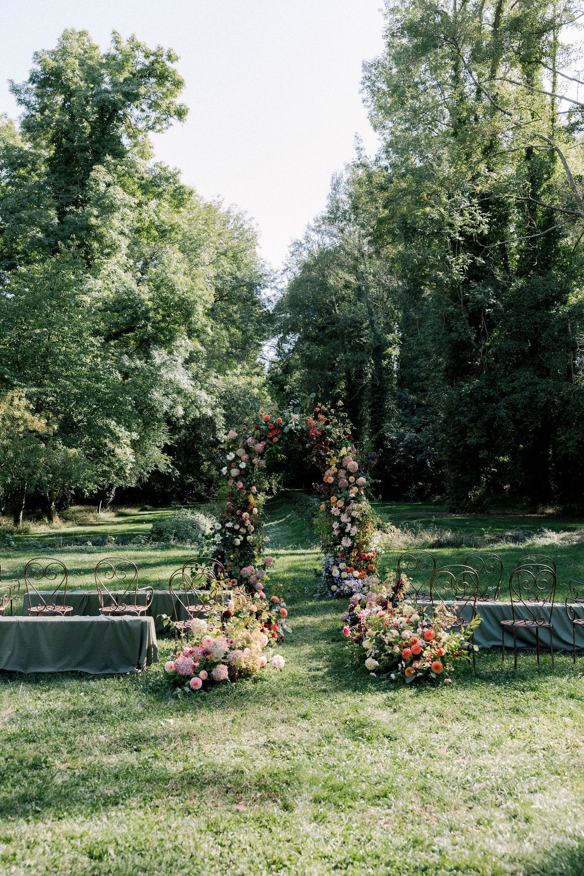 Two tall floral arrangements stand on a lawn between trees, with low hedged beds in the background.