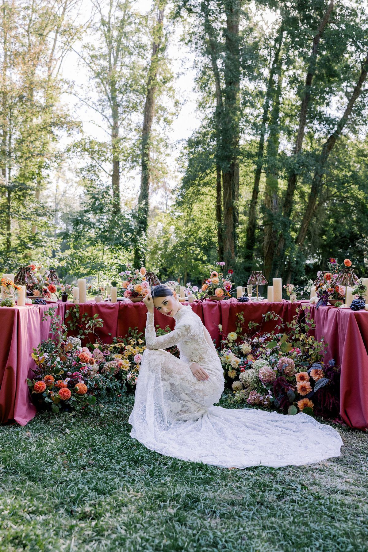 The bride in a long-sleeved lace gown with a long train crouches on the grass in front of a burgundy table with colourful flowers.